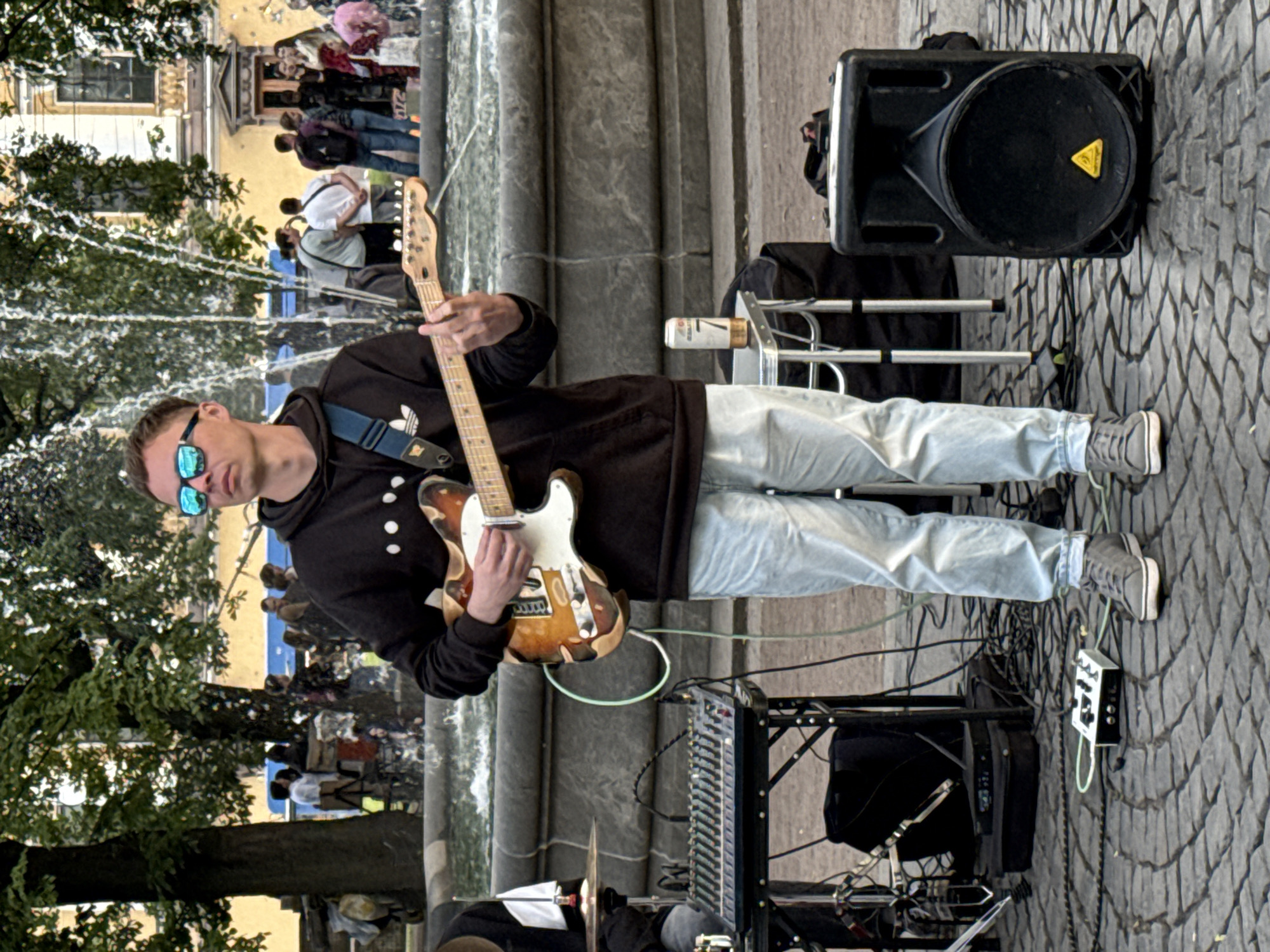 A man plays a Fender Telecaster electric guitar outdoors, possibly as a street performer. He wears a black hooded sweatshirt with white dots and light blue jeans, along with blue mirrored sunglasses. The guitar has a sunburst finish. Behind him, a large black speaker with a yellow triangular logo sits on the cobblestone ground. Various cables and a mixing board are visible, suggesting a sound setup for a performance. In the background, a crowd of people is gathered, some appearing to be tourists, with trees and a building with ornate architecture. The scene is set in Saint Petersburg, Russia, and the lighting suggests it is daytime. The overall mood seems casual and lively, with the man focused on his music.