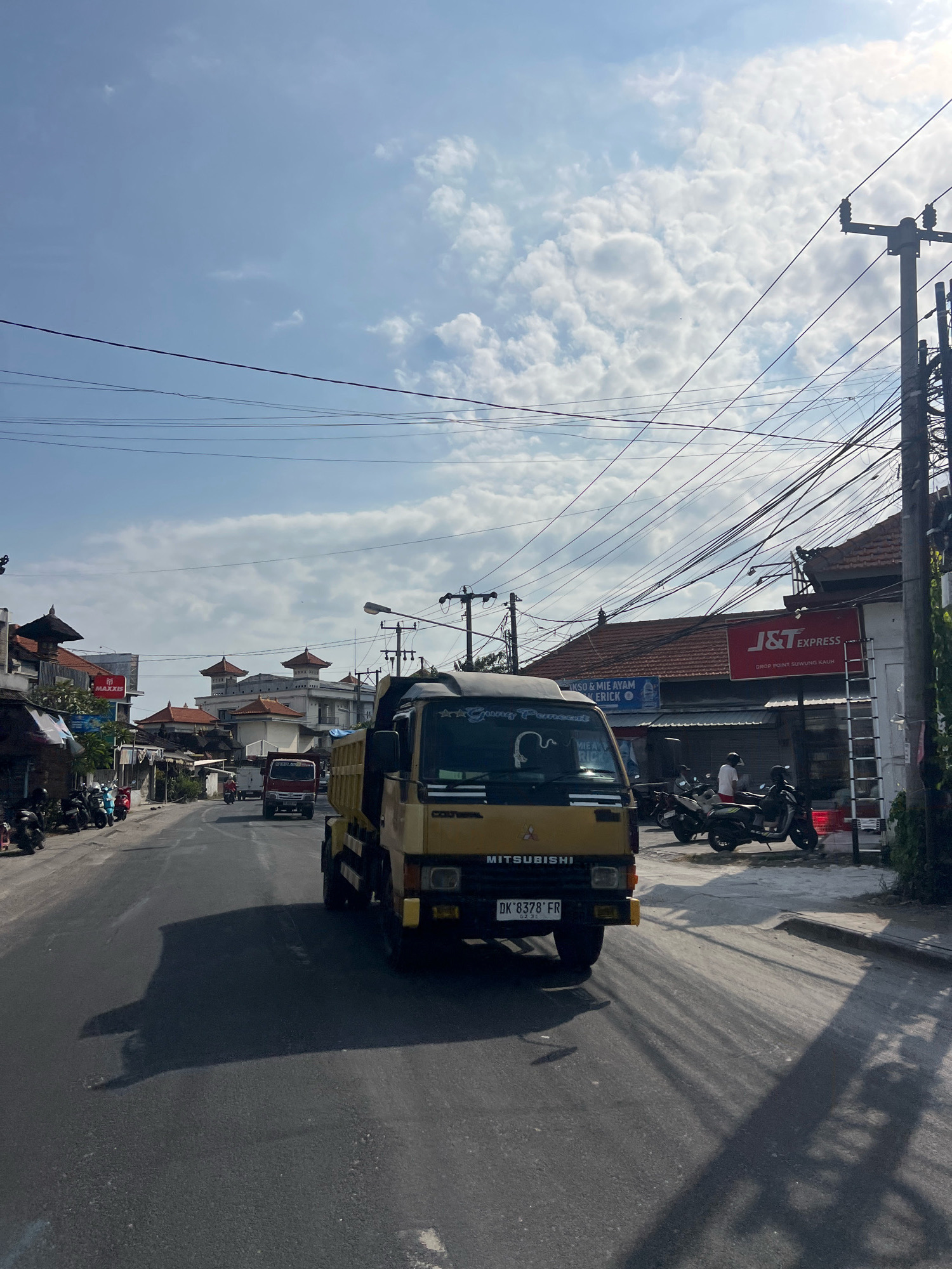 A yellow Mitsubishi dump truck drives down a paved street in Kuta, Indonesia. The truck is in the foreground, casting a long shadow on the asphalt. The street is lined with buildings, some with traditional Balinese architecture featuring tiered roofs. Several motorcycles are parked along the side of the road. In the background, a red truck is also visible. The sky is a bright blue with scattered white clouds, and electrical wires crisscross overhead. A sign for "J&T Express" is visible on one of the buildings. The scene appears to be a typical day in a bustling commercial area.