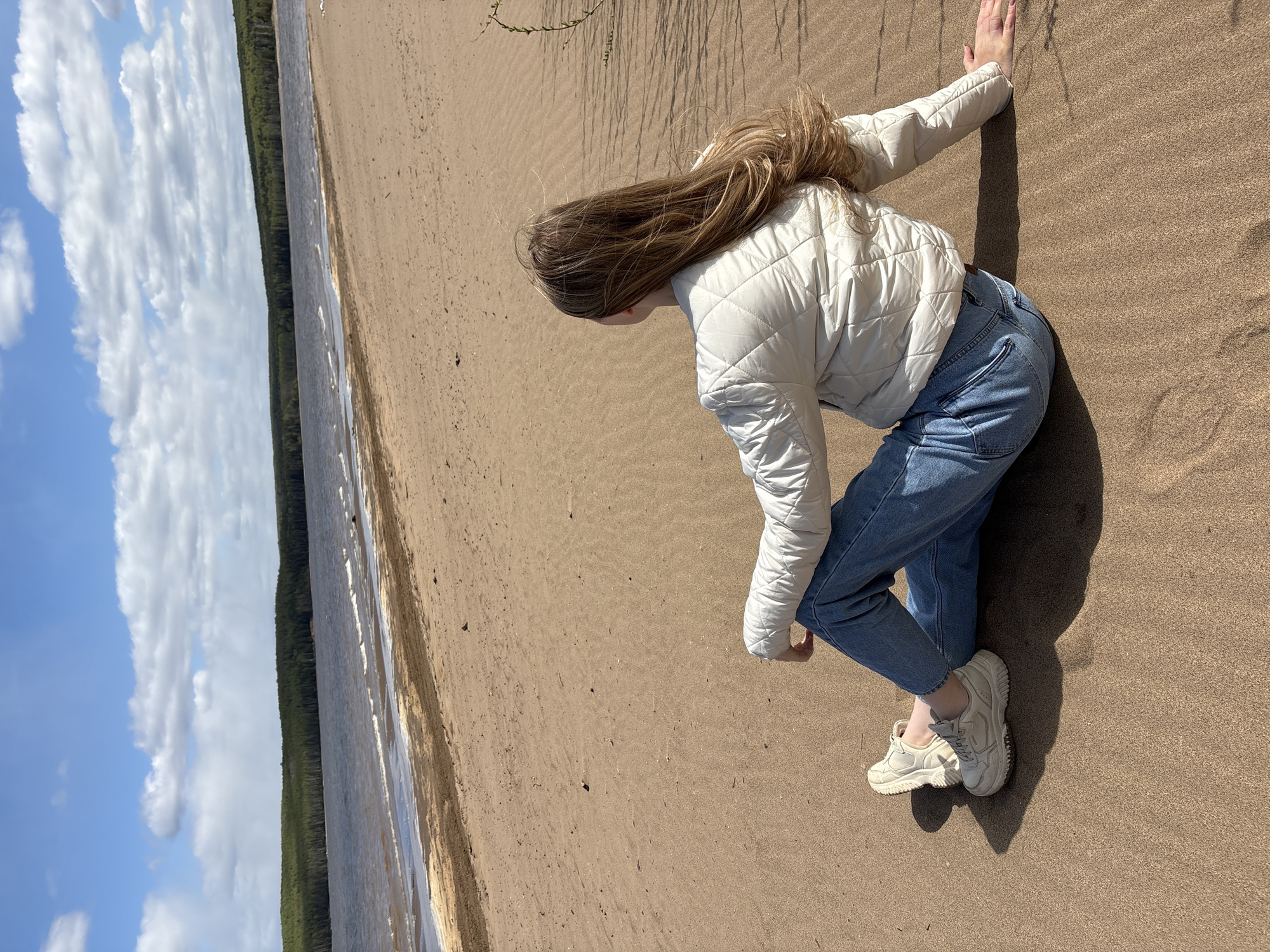 A woman in a white jacket, blue jeans, and white sneakers is positioned on a sandy surface next to a body of water. Her blonde hair is visible as she leans forward. The shoreline transitions from sand to water, with a line of trees visible on the opposite bank. The sky is partially cloudy. The location is likely Chaikovsky, Russia.
