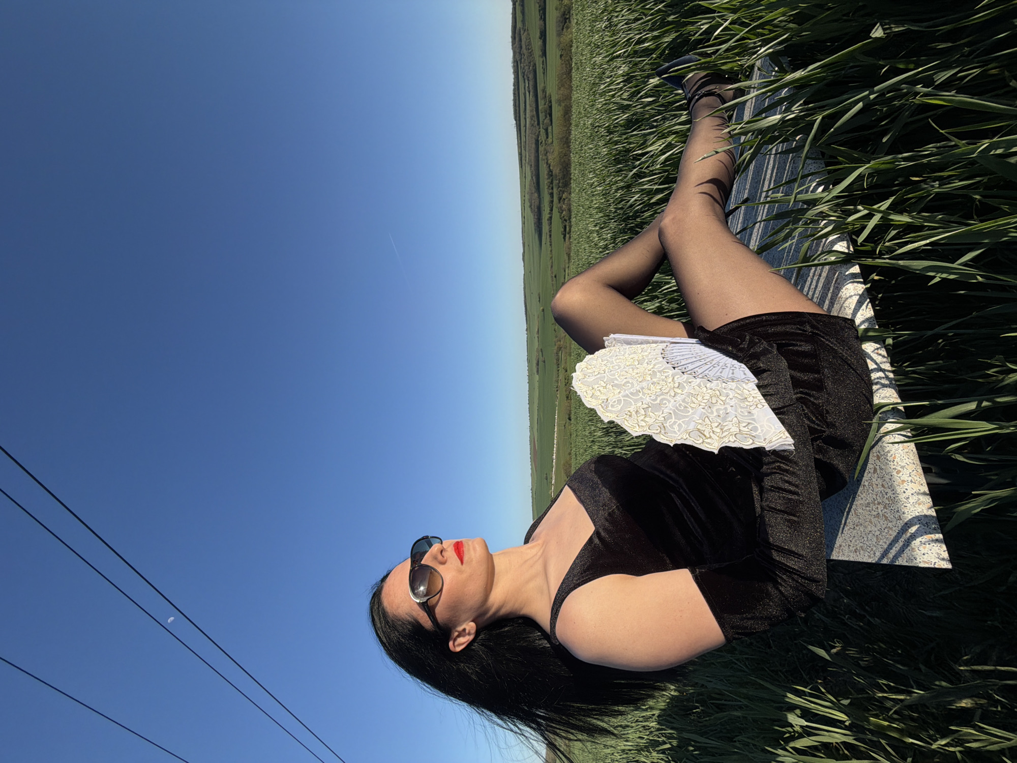 A woman is reclining on a flat, likely stone surface amidst tall, green grass. She is wearing a black dress, black tights, and heeled shoes. She has sunglasses on and is holding a white lace fan. The sky above is a clear, deep blue. A landscape stretches across the background, featuring a green field and trees. Power lines are visible in the sky. The location is near Pleven, Bulgaria.