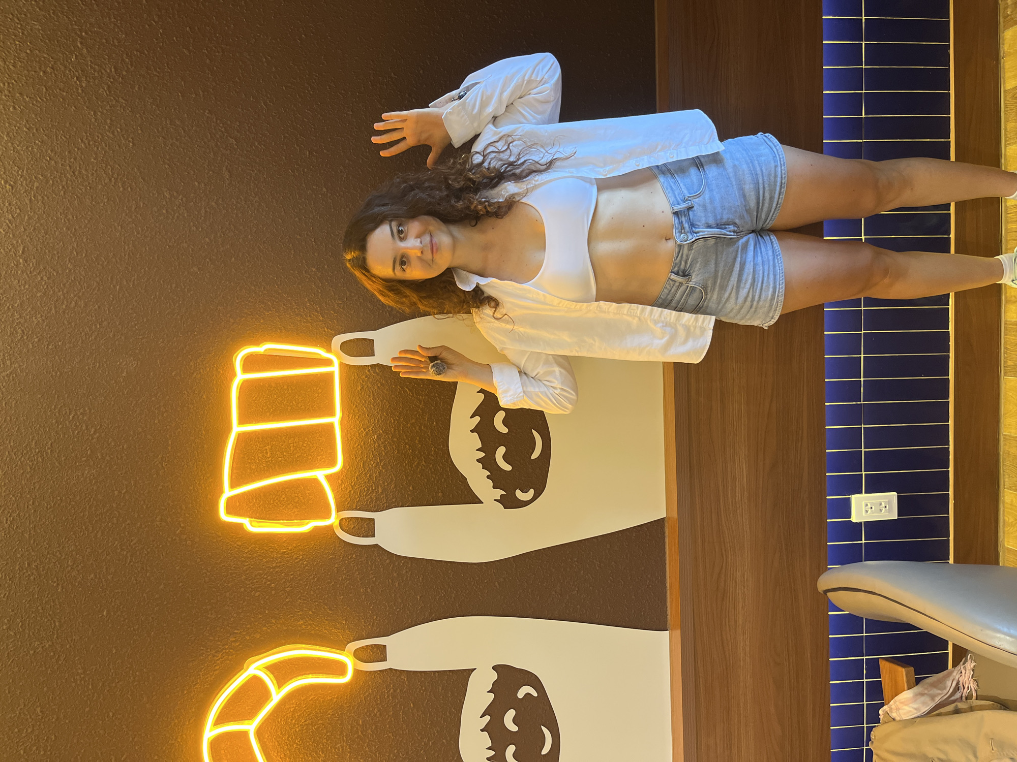 A woman stands facing forward in a room with a dark brown wall adorned with neon orange art depicting two hands holding croissants. The woman is wearing a white crop top, an unbuttoned white shirt, and denim shorts. Her arms are raised with her palms facing forward, and she is wearing rings on her fingers. To the left of the woman, a portion of a light gray chair is visible. In the background, beneath the wall art, is a wooden ledge and then a wall covered in vertically arranged dark blue tiles. The floor appears to be yellow. The location is identified as Phan Thiết, Vietnam, within a commercial establishment, likely a cafe or bakery, given the croissant art.