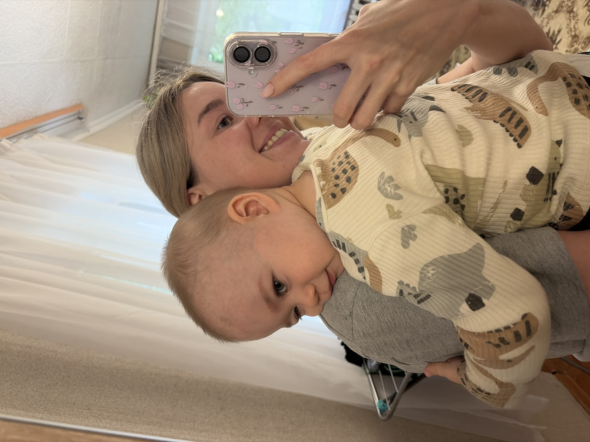 A person is taking a selfie with a baby. The person is holding a smartphone with a pink floral case. The baby is wearing a light-colored patterned top. They appear to be indoors. No specific location in the city of Chaikovsky, Russia, can be determined from the image.