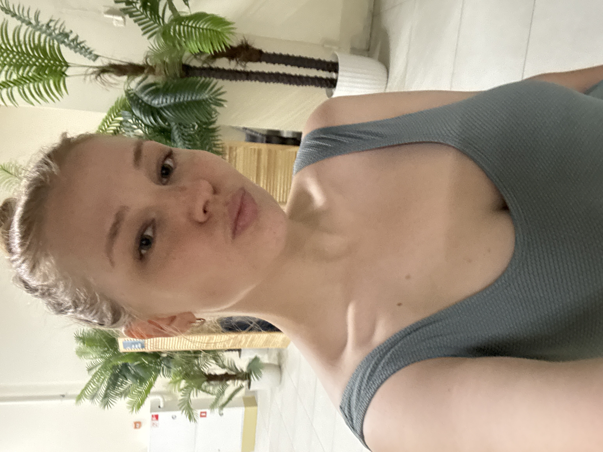 A young woman with blonde hair pulled back is taking a selfie in an indoor setting. She is wearing a grey, ribbed tank top. Her lips are pursed, and she is looking at the camera. Behind her are several large artificial palm plants in white pots. To the left of the frame, there is a white cabinet with red and white labels. The floor appears to be tiled in a light color. The background suggests an indoor public space, possibly a mall, hotel lobby, or office building. The image does not contain any information that allows for pinpointing the location to Yaroslavl, Russia.
