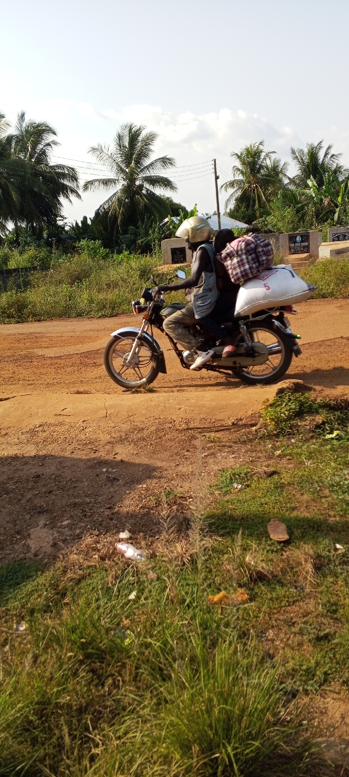 A dark-colored motorcycle carrying two individuals and two large items of luggage travels along a reddish-brown dirt road in Dzodze, Ghana. The driver, a male wearing a light-colored vest, camouflage pants, white sneakers, and a gold-colored full-face helmet, operates the motorcycle. A second, smaller individual, likely a passenger, sits behind the driver, wearing a plaid shirt and pinkish footwear; their upper body is largely obscured by a plaid fabric bundle. A large white sack marked with a red "5" is secured to the motorcycle's rear rack.
The immediate foreground consists of reddish-brown dirt and sparse green grass, with discarded litter including a white plastic bottle. The background features abundant green tropical vegetation, dominated by tall palm trees, under a light blue sky with scattered white clouds. A utility pole stands on the right side of the road, and several light-colored stone structures, consistent with gravestones or tombs, are visible in the mid-background, indicating proximity to a cemetery.