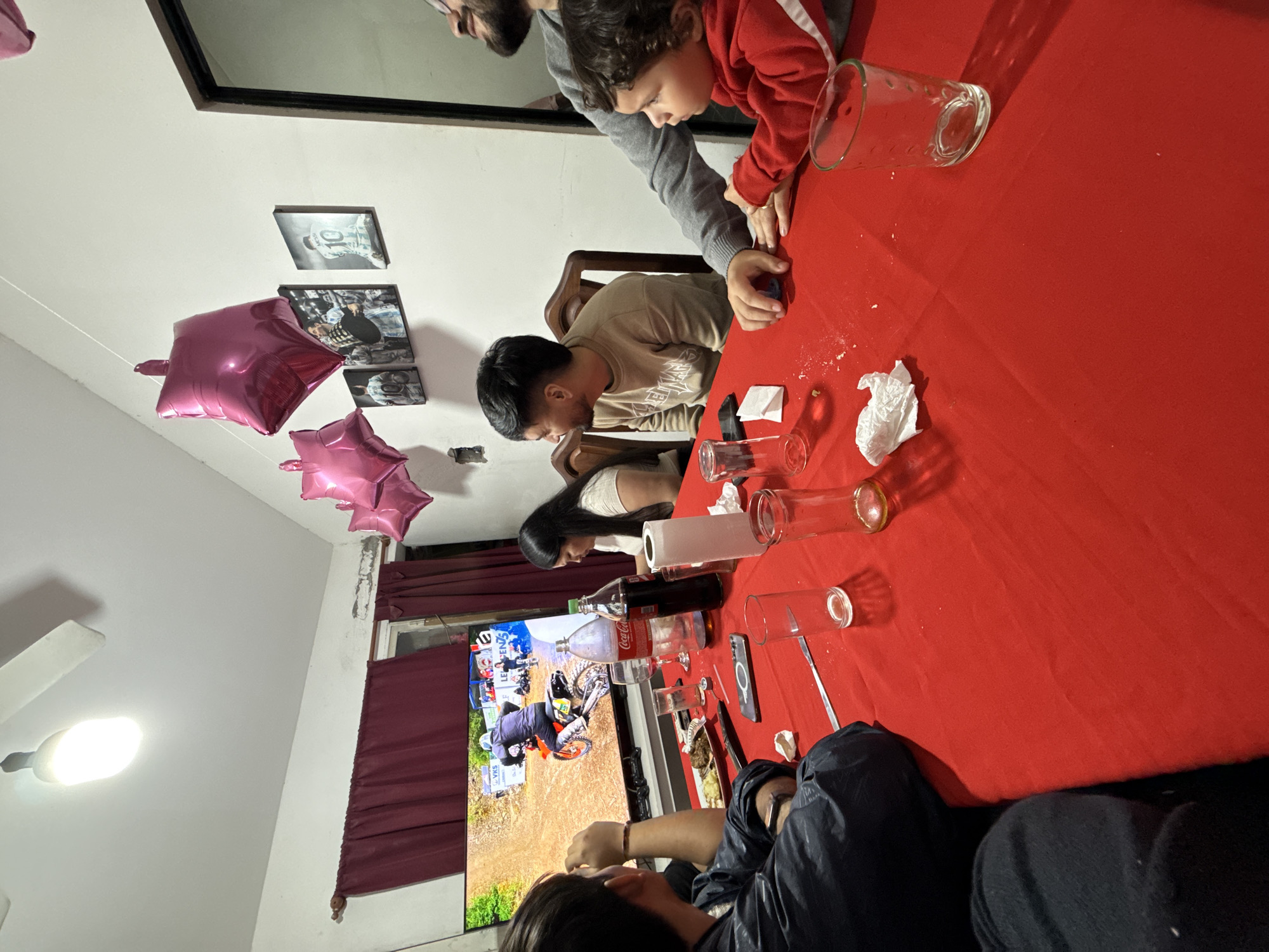 A family is gathered around a table covered with a red tablecloth. Several clear glass cylinders or drinking glasses are scattered across the table. Some people are sitting and watching TV. On the wall are photos in black frames and two pink star-shaped balloons. A Coca-Cola bottle is on the table. There are maroon curtains in front of a window. The location is in Santa Rosa, Argentina, based on the image's visual cues like the type of beverage on the table.