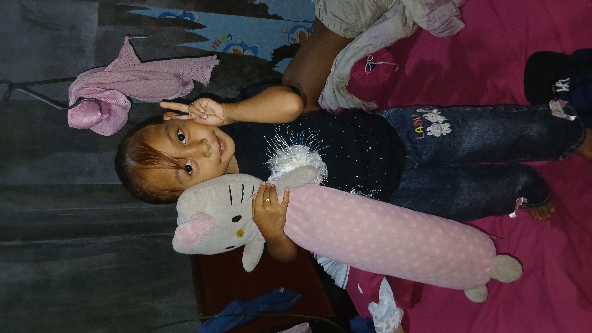 A young girl stands in what appears to be a bedroom, holding a large stuffed Hello Kitty toy and a pink polka-dotted bolster pillow. She is wearing a black t-shirt with rhinestones and blue jeans with patches of two white bunnies and the word "LABU". The girl is making a peace sign with her right hand. The room has a dark, textured wall in the background, with a pink scarf hanging from a hook. A blue tarp with cartoon characters, possibly Doraemon, is visible on the wall. The girl is standing on a bed with a pink sheet. The scene is dimly lit, suggesting an indoor environment. The location is Keudejungkagajah, Indonesia.
