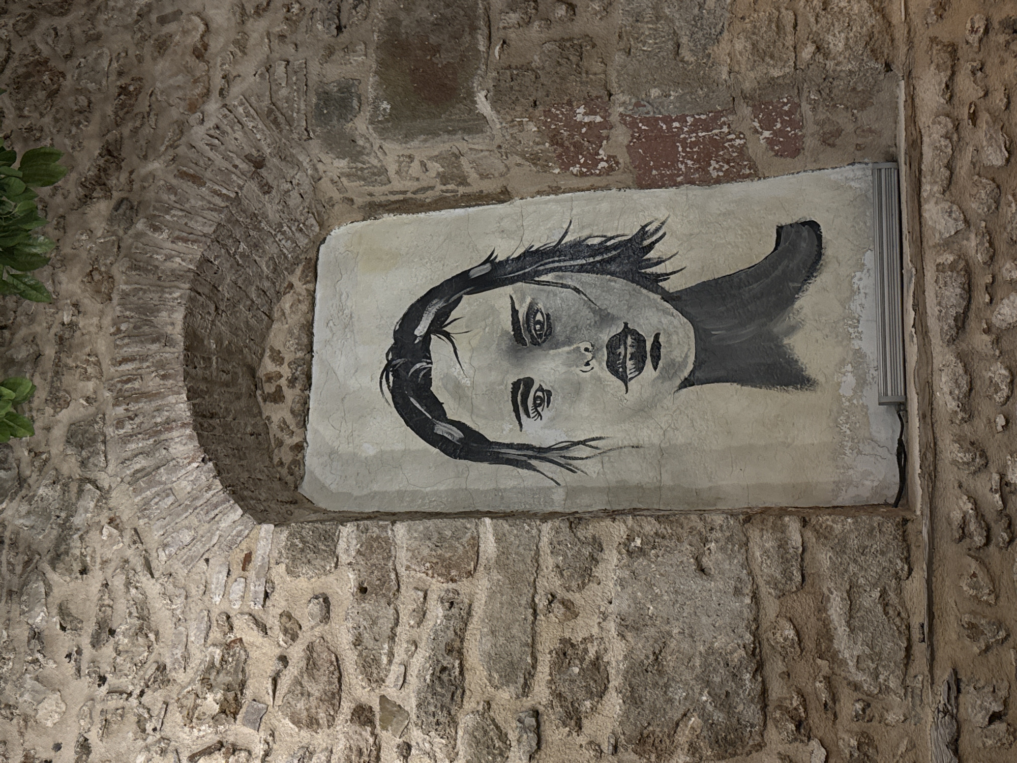 A black and white painting of a woman's face is affixed to a rough-hewn stone wall in Antalya, Turkey. The painting is on a vertical rectangular surface, appearing to be a panel or canvas, set within an arched recess in the stone. The woman's hair is dark and styled to frame her face, with some strands appearing windswept. Her eyes are detailed with prominent lashes, and her lips are full and slightly parted. The painting utilizes broad strokes, particularly for the hair and neck area, suggesting a stylized or impressionistic approach. The stone wall surrounding the artwork is constructed from irregular, varying-sized stones with visible mortar. Above the recess, the stone forms a rounded arch. Green leaves are visible at the top edge of the image, hanging down over the stone structure. The bottom of the painting rests on a narrow ledge. The overall scene suggests an outdoor, possibly historic, urban setting in Antalya.