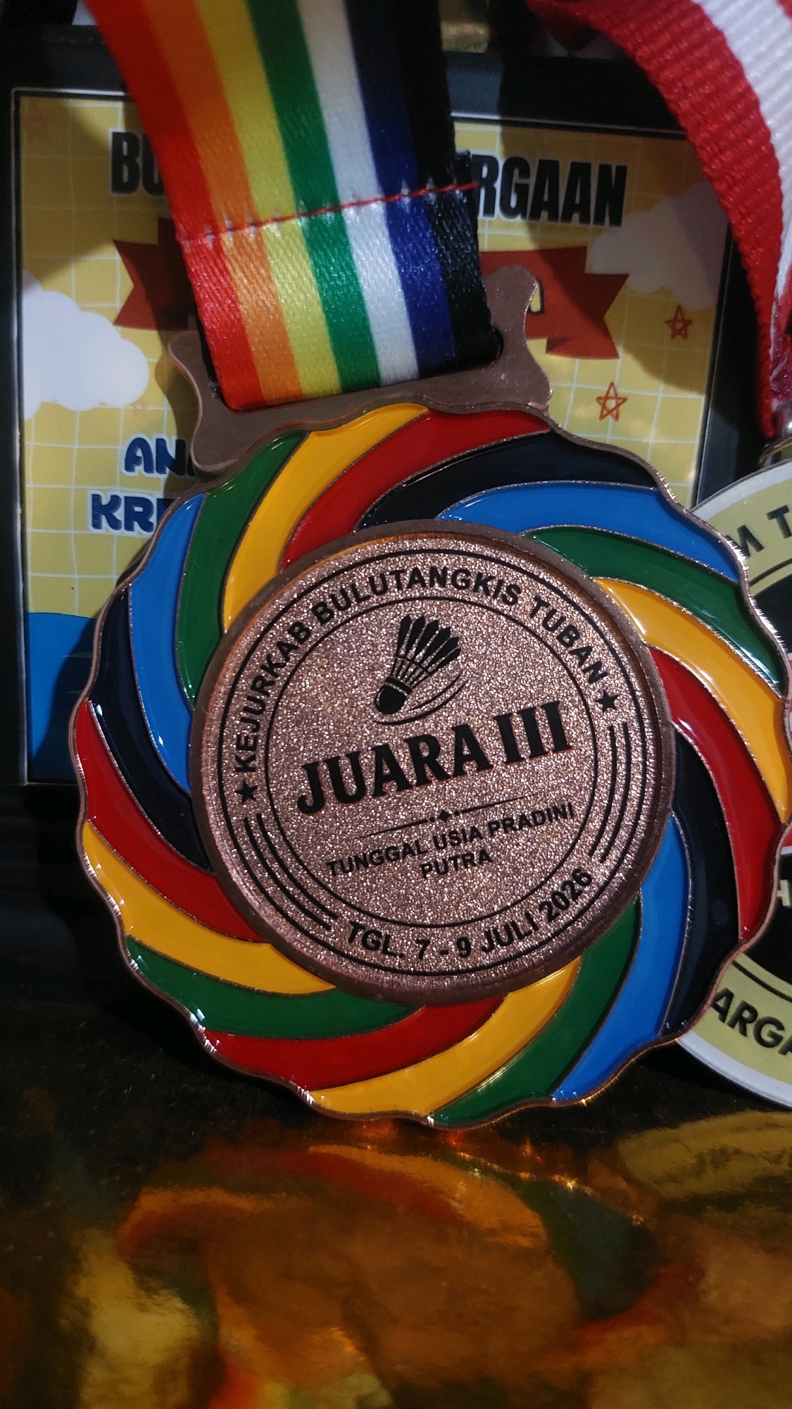 The image shows a close-up of a medal with a rainbow-colored ribbon. The medal is bronze-colored and has an embossed design featuring a badminton shuttlecock and text in Indonesian. The text reads "KEJURKAB BULUTANGKIS TUBAN" around the top, "JUARA III" in large letters in the center, and "TUNGGAL USIA PRADINI PUTRA" below that. At the bottom, it says "TGL. 7 - 9 JULI 2025". The medal is set against a blurred background that appears to be some kind of award certificate or backdrop with the words "BU.. RGAAAN" visible. The medal is resting on a reflective gold surface, which may be part of the display or the medal itself. The image does not provide any information about the location in Jatirogo, Indonesia.