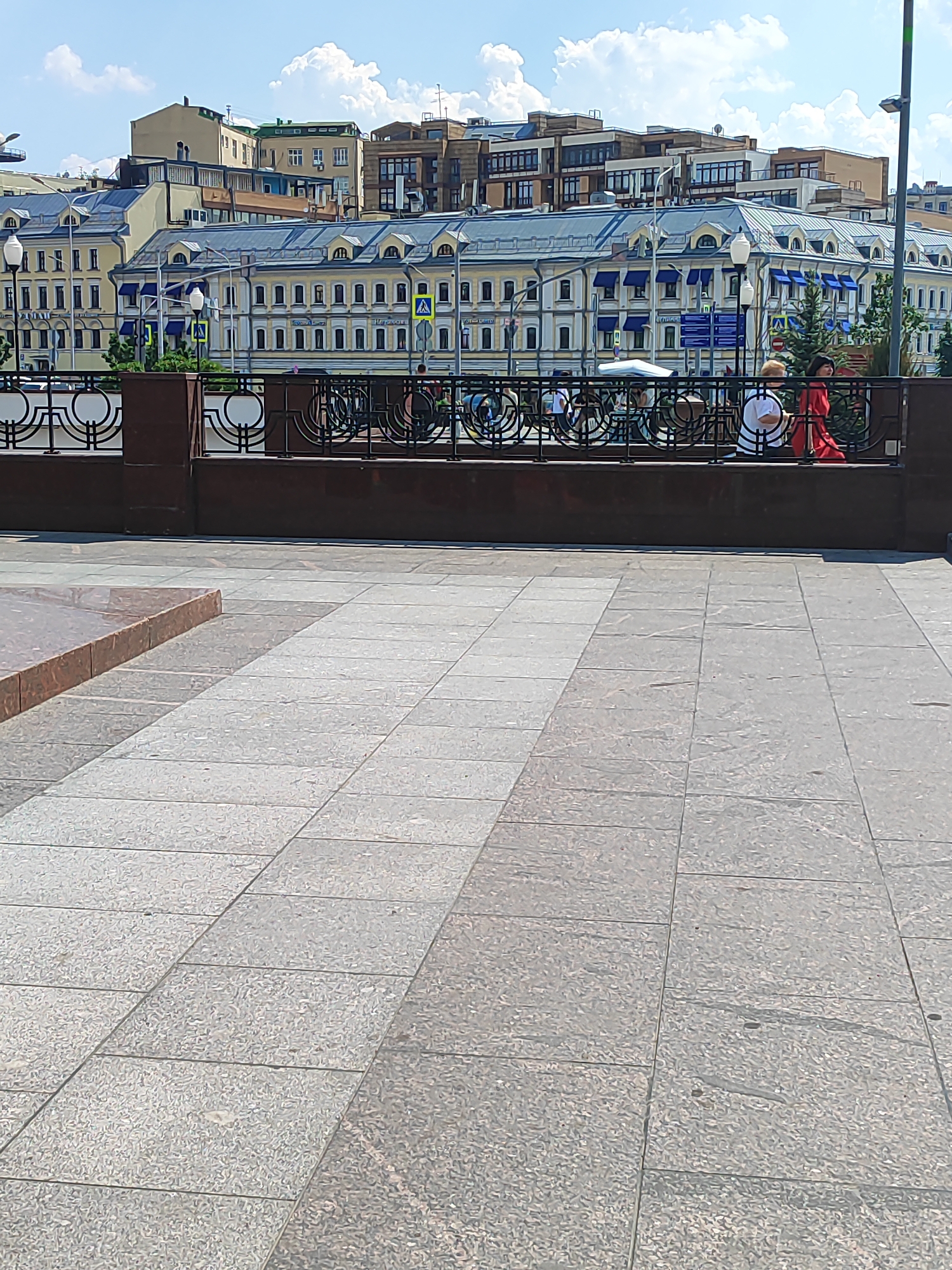 The image shows a view of a promenade area in Moscow, Russia, with a stone-tiled walkway in the foreground and a row of buildings in the background. The buildings are a mix of architectural styles, with some appearing older and more ornate, while others are more modern. A decorative metal fence separates the promenade from the street and the buildings. Two women are visible behind the fence, one wearing a red dress and the other in a white top. The sky is blue with scattered white clouds. The overall scene suggests a pleasant day in an urban setting.