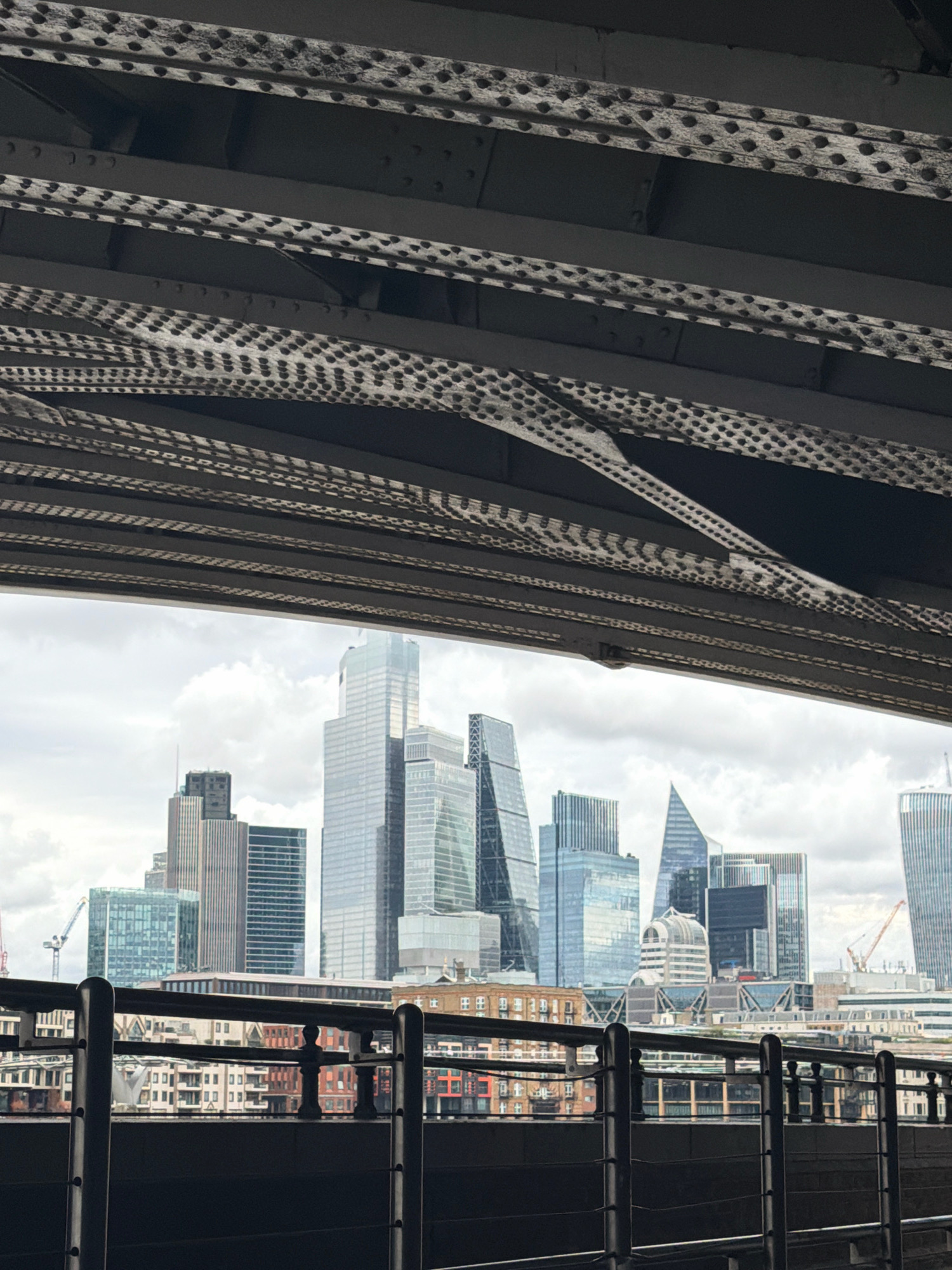 This is a photograph taken from underneath a bridge, looking out at the London skyline. The foreground is dominated by the dark, industrial metal structure of the bridge, with its rivets and supporting beams clearly visible. Through the gap formed by the bridge, a view of the city's financial district unfolds.
The skyline features a collection of modern skyscrapers, characterized by their glass facades and varied architectural designs. Notable buildings include those with sharp, angular shapes and others with more traditional rectangular forms. In the middle ground, there are lower-rise buildings, some appearing to be older structures with brickwork and rows of windows, interspersed with more modern constructions.
On the left side of the skyline, a construction crane is visible, indicating ongoing development in the area. The sky is overcast with clouds, suggesting a typical temperate London day. The overall scene captures the contrast between the man-made industrial infrastructure of the bridge and the modern urban landscape of London. There are no people visible in the image.