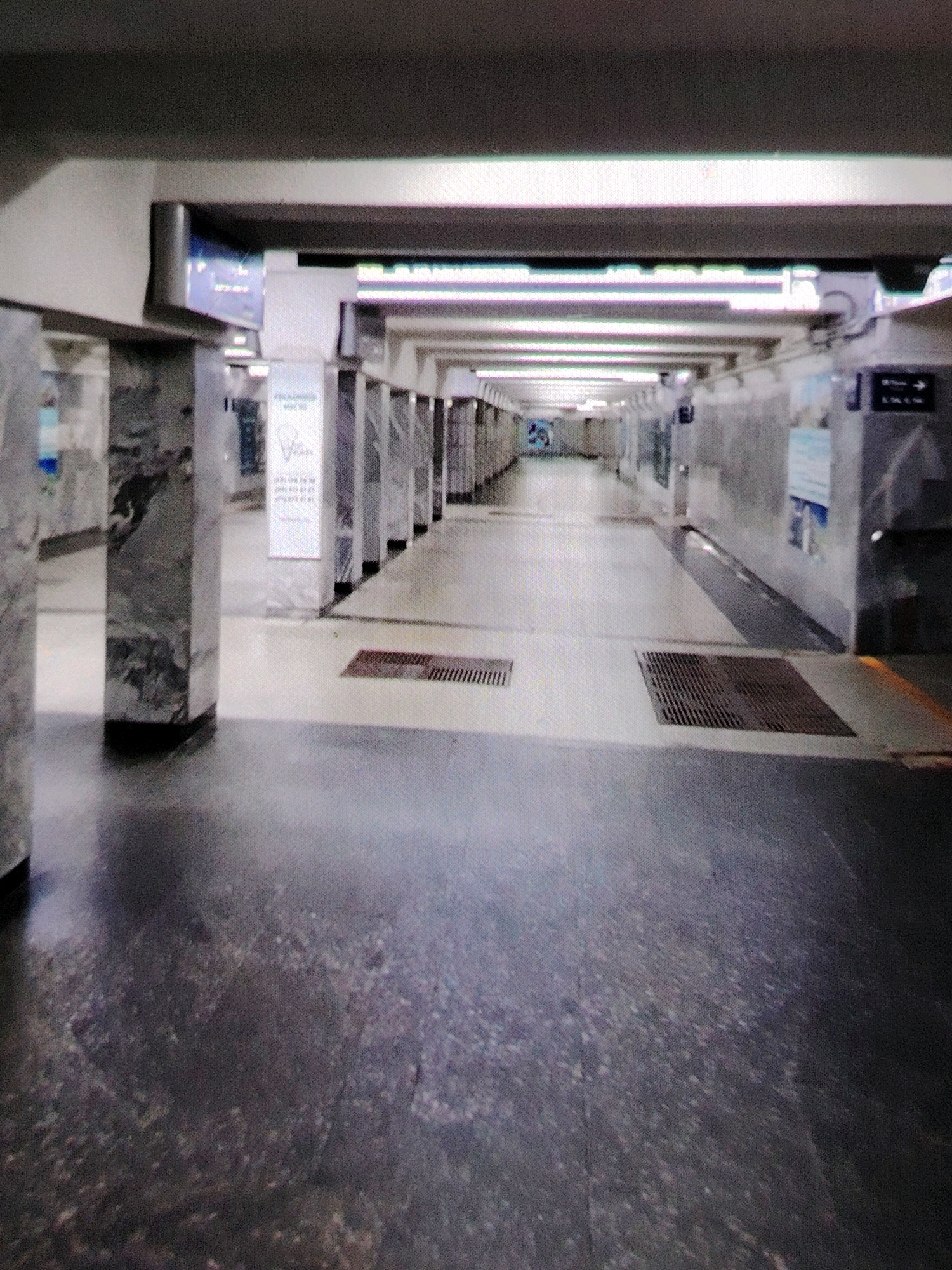 An overhead view of a long, empty subway station corridor in Mazyr, Belarus. The station features marble-clad columns, fluorescent lighting, and a polished concrete floor. A sign with text and an image is visible on one of the columns. The corridor stretches into the distance, with no people or activity apparent.