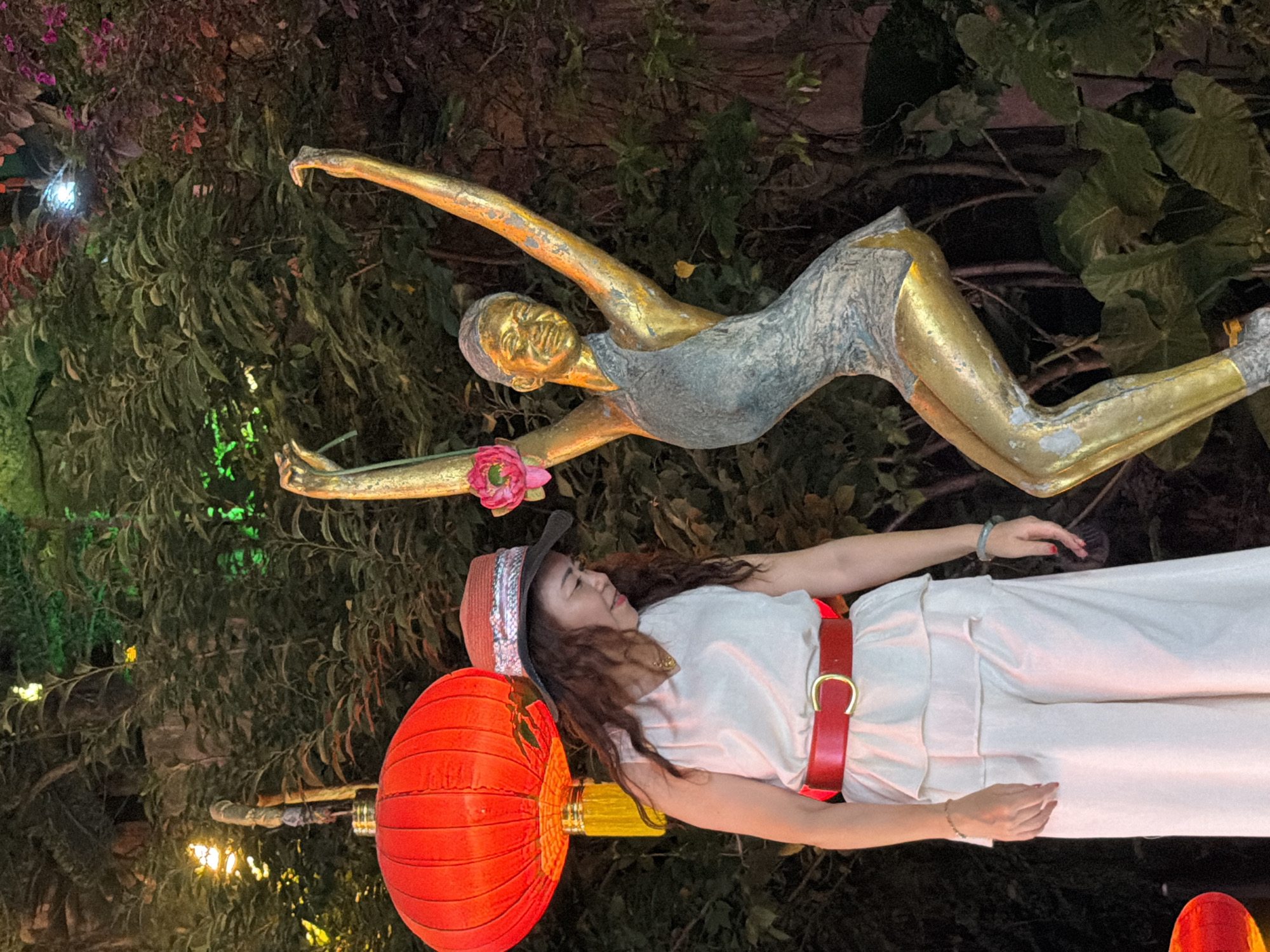 The image depicts a woman standing next to a golden statue of a dancer. The woman is wearing a white sleeveless top, a wide red belt with a gold buckle, and loose-fitting white pants. She has long, wavy brown hair and is wearing a brown hat with a silver band. She is looking downwards, with her hands clasped in front of her. The statue is of a slender female figure in a dynamic dancing pose, with its arms raised and one leg extended. The statue appears to be made of a material that is partially golden and partially a greenish-blue hue, suggesting age or a specific artistic finish. A pink flower is held in the statue's left hand. To the left of the woman is a large, bright red Chinese lantern hanging from a pole, with a yellow fringe. The background consists of dense, dark green foliage, with some areas illuminated by colored lights, including green and possibly pinkish-red, creating an atmospheric, possibly festive, environment. The scene is likely outdoors, possibly in a garden or a park during an evening event. Without further information, it is not possible to definitively confirm the location as Yuzhong District, China, but the presence of the Chinese lantern suggests an East Asian setting.