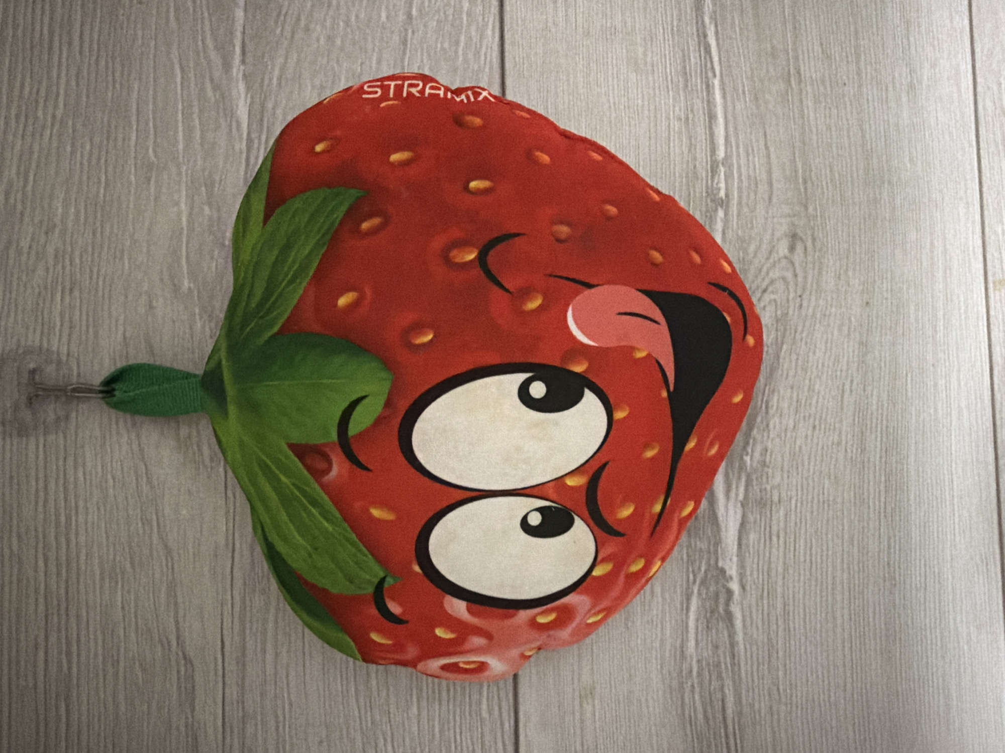 A plush strawberry toy with a smiling face and large cartoon eyes is hanging from a hook against a light gray wooden plank wall. The toy is red with yellow seeds and green leaves at the top. Text reading "STRAMIX" is visible vertically on the right side of the toy. The background appears to be an indoor wall with a wood grain texture. There are no people or animals present in the image. The scene is static and does not depict any action or interaction. The location is not specified as Semey, Kazakhstan.