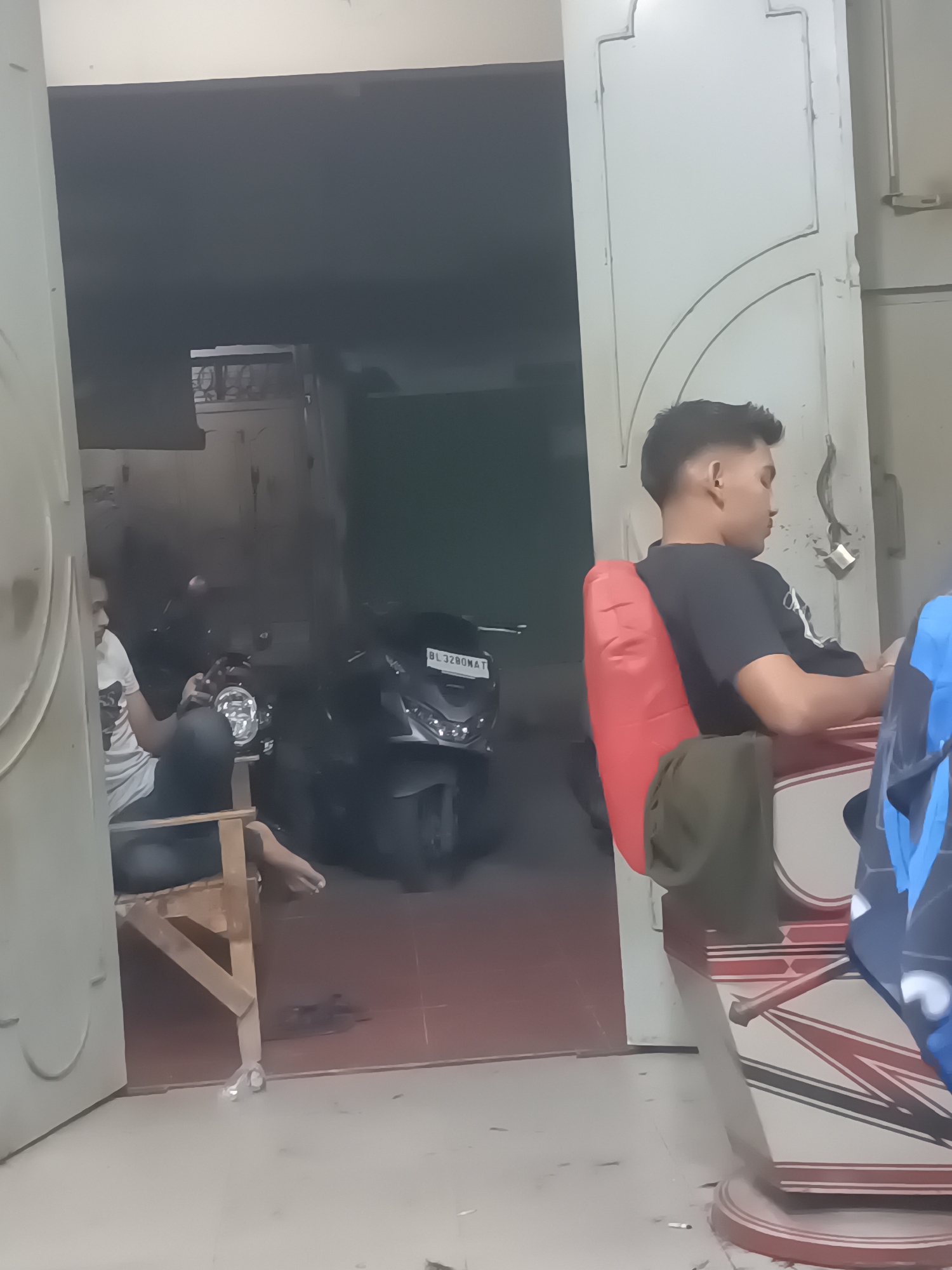 In Lhokseumawe, Indonesia, a man with short, dark hair is sitting in a barber's chair, looking down and to his right, his facial features are not clearly visible. He is wearing a dark t-shirt with white accents and a red cushion is behind his back, with a green cloth draped over it. The barber's chair has a patterned base and is upholstered in red and white. To the left of the barber's chair, a second man, wearing a white t-shirt and dark pants, is seated on a wooden stool, looking at his phone and holding it in his right hand. He is positioned in front of an open doorway. Behind him, partially visible through the doorway, is a dark-colored motorcycle with a license plate reading "BL 3280 NAT". The floor appears to be tiled in a reddish-brown color. The overall lighting is dim, suggesting an indoor setting, possibly a barber shop or a similar establishment.