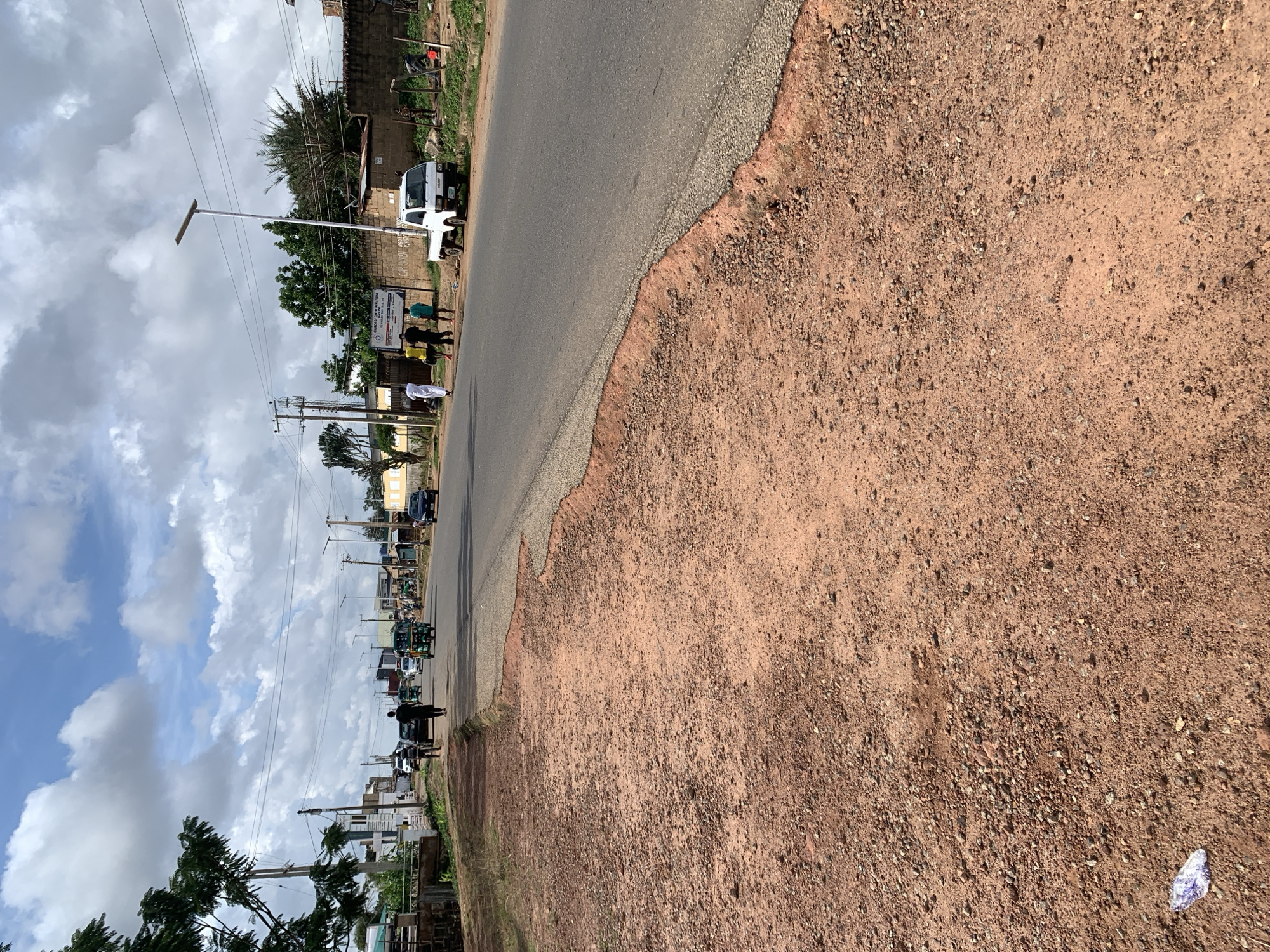 The image shows a street scene in an outdoor urban environment under a cloudy sky. In the foreground, a rough, reddish-brown dirt and gravel surface slopes down to meet the asphalt road. The road itself is paved and appears to be a two-lane street with vehicles in motion. Several people are walking on the sidewalk or near the edge of the road. Traffic includes a green auto-rickshaw (tricycle taxi), a white utility truck, and other cars further down the road. Buildings and trees line the side of the road, some with fences or walls. Electrical poles and wires are visible. In the distance, the sky is filled with large, white cumulus clouds. The overall impression is a typical street view in a warm, possibly tropical, climate.