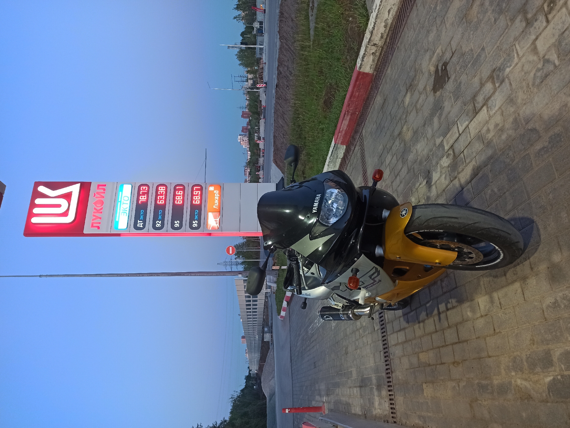 Here is a description of the image:
A Yamaha motorcycle sits parked on a brick surface in front of a Lukoil gas station. The prices on the gas station sign are displayed as follows: ДТ ЭКТО 78.73, 92 ЭКТО 63.38, 95 68.61, 95 ЭКТО 68.97, and "Ликард". The motorcycle is a black and silver color scheme, with a gold front fender and Yamaha logo on the fairing. A single exhaust pipe is visible. The background shows a clear blue sky transitioning to a hazy horizon. Other buildings and foliage line the area near the road in the distance. The location appears to be in the city of Unknown, Unknown.