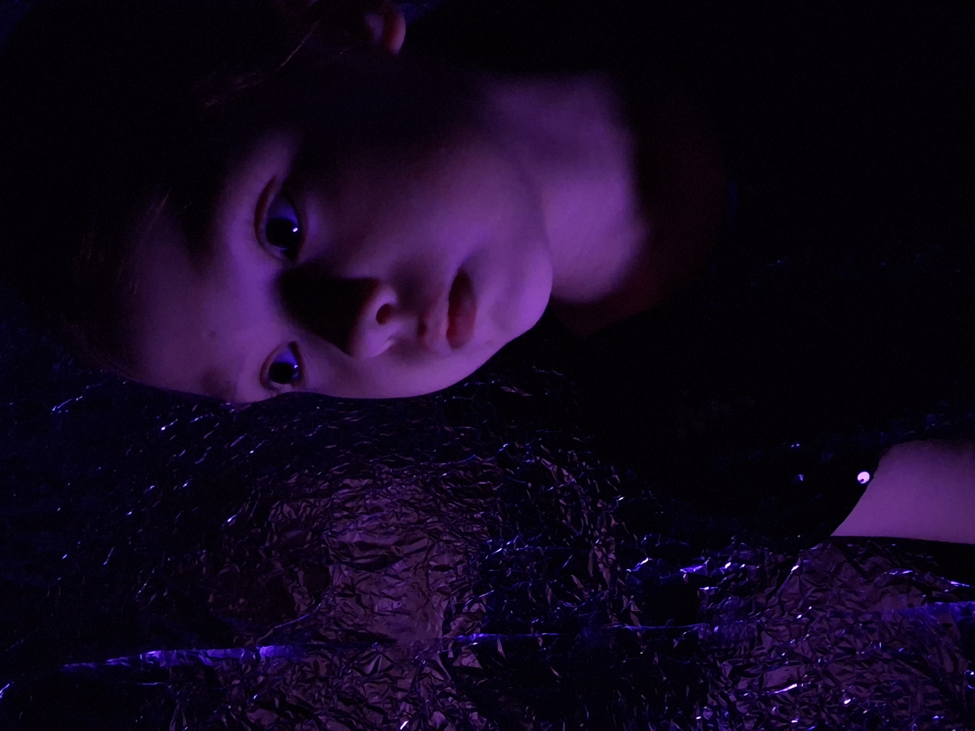 A young child is lying on a crumpled, reflective surface, possibly aluminum foil. The child's head is positioned in the upper left of the frame, with their eyes looking toward the viewer. The child is illuminated by a purple light source. The location of the photograph is Chaikovsky, Russia.