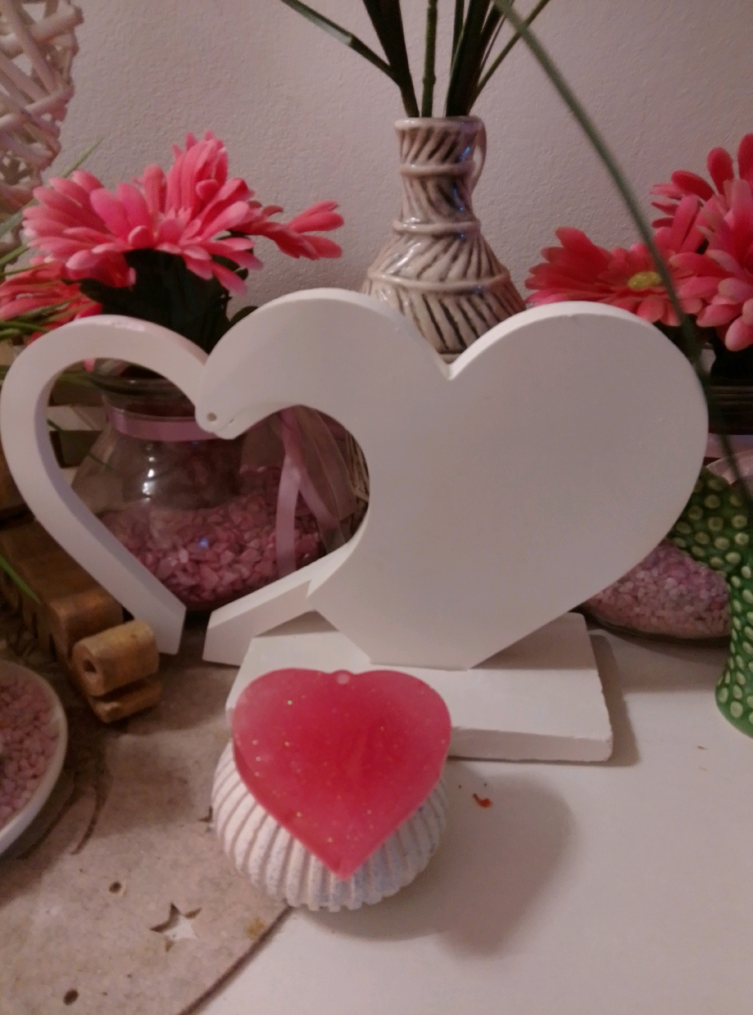 A still life photograph is presented, featuring a collection of decorative items with a Valentine's Day theme. In the foreground, a heart-shaped red soap or wax ornament with glitter rests on a ridged, cream-colored base. Behind it, a large, white, hollow heart-shaped wooden or plaster sculpture is prominently displayed, appearing to be a stand or centerpiece. This sculpture is positioned in front of a glass jar filled with pink decorative stones or potpourri, tied with a pink ribbon.
To the left of the sculpture, a rustic wooden object, possibly part of a craft or display, is visible. To the right, behind the white heart, are several vibrant pink artificial gerbera daisies, some partially obscured. A tall, narrow, textured vase with a beige and brown striped pattern stands behind the flowers and the white heart, holding what appear to be green plant stems. The background is a plain, light-colored wall. The overall setting suggests an indoor decoration, likely in a home or shop, with a romantic or festive aesthetic. The image does not provide any information that would allow for its location to be identified within the city of Wedel, Germany.
