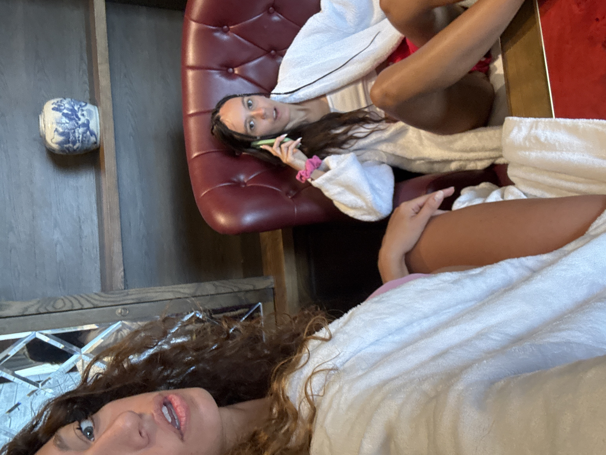 Two women are in a room. One woman is lying on a red leather couch and holding a green phone to her ear. She is wearing a white robe. The other woman is partially visible in the foreground wearing a white robe. A blue and white ceramic vase is on a shelf. The location is not evident from the image.