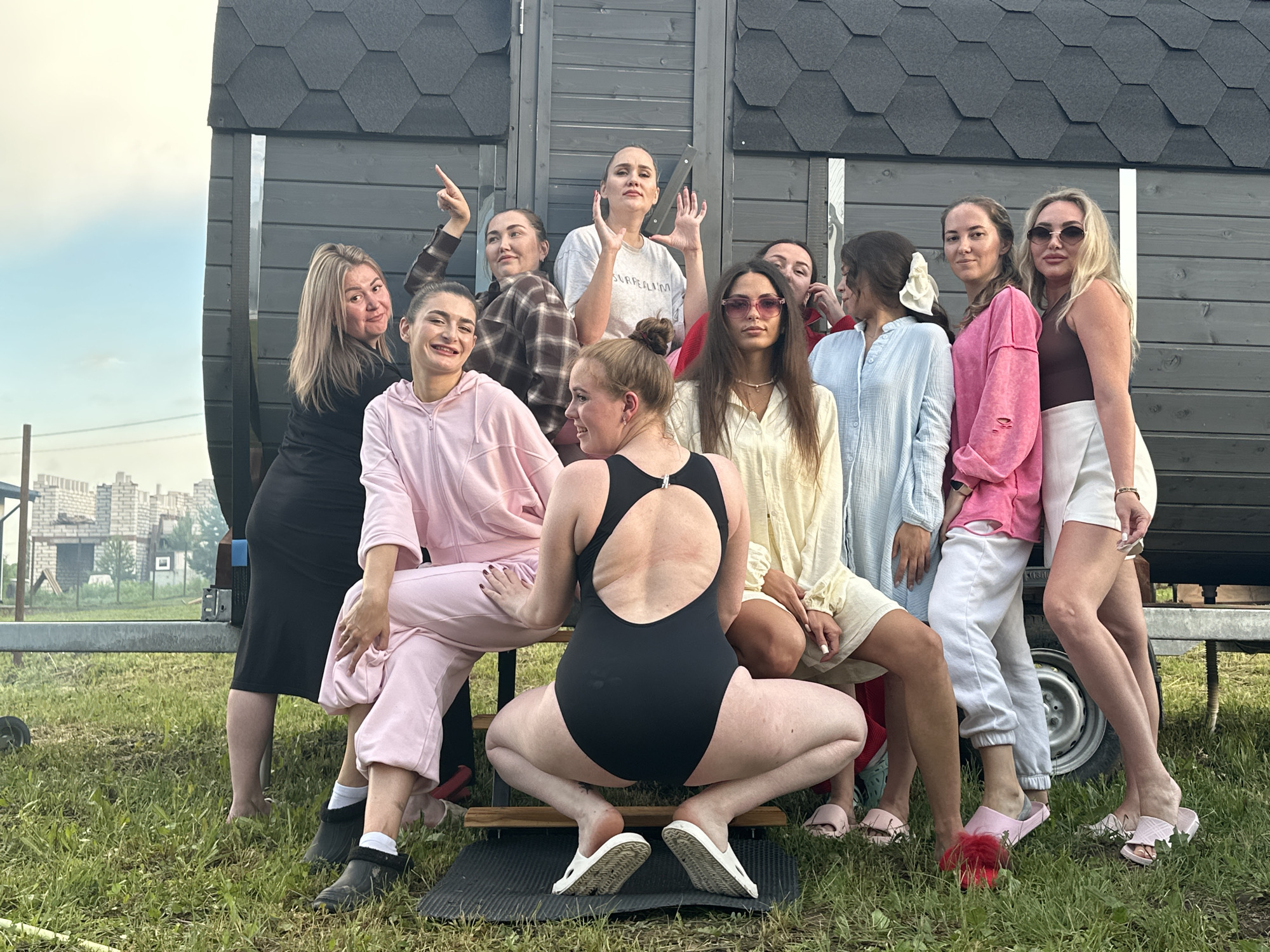 In Zav'yalovo, Russia, a group of ten young women are gathered outside a dark wooden structure that appears to be a mobile sauna. The structure is situated on a grassy area, with some construction visible in the background. The women are dressed in various casual and athletic wear, including sweatpants, hoodies, a swimsuit, and dresses. Several of them are posing for the camera, with some smiling, pointing, or making gestures. The overall atmosphere appears to be relaxed and social.