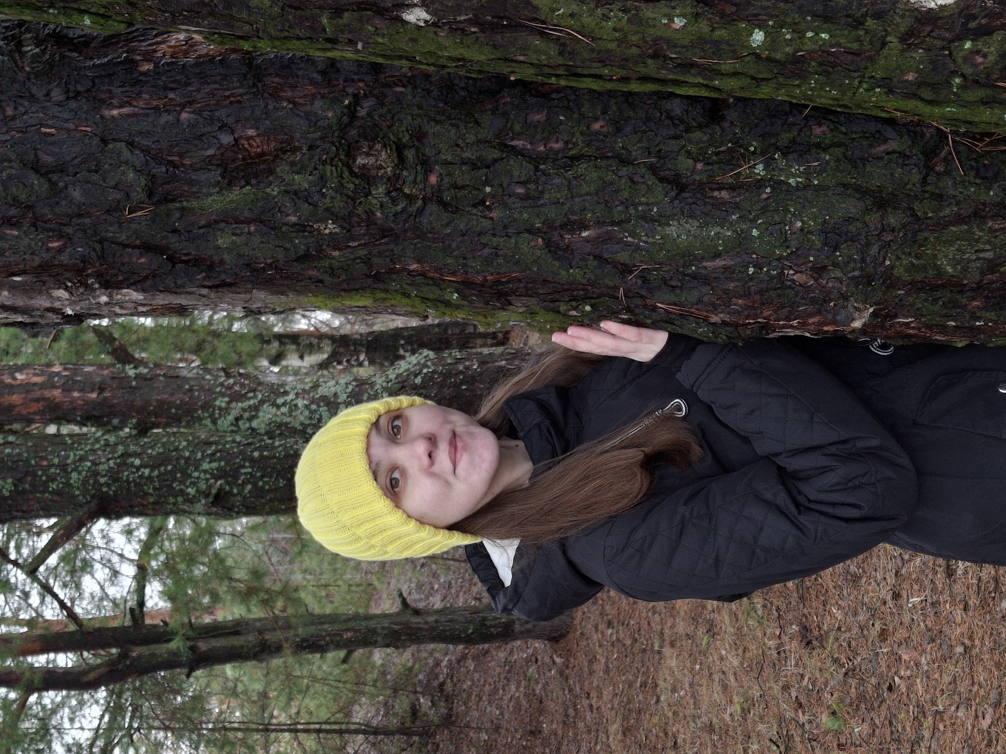 A woman in a yellow knit cap and black jacket stands between two trees in a forest. The tree trunks are covered with moss. The ground is covered with brown pine needles and fallen leaves. The location is likely in a wooded area of Chaikovsky, Russia.