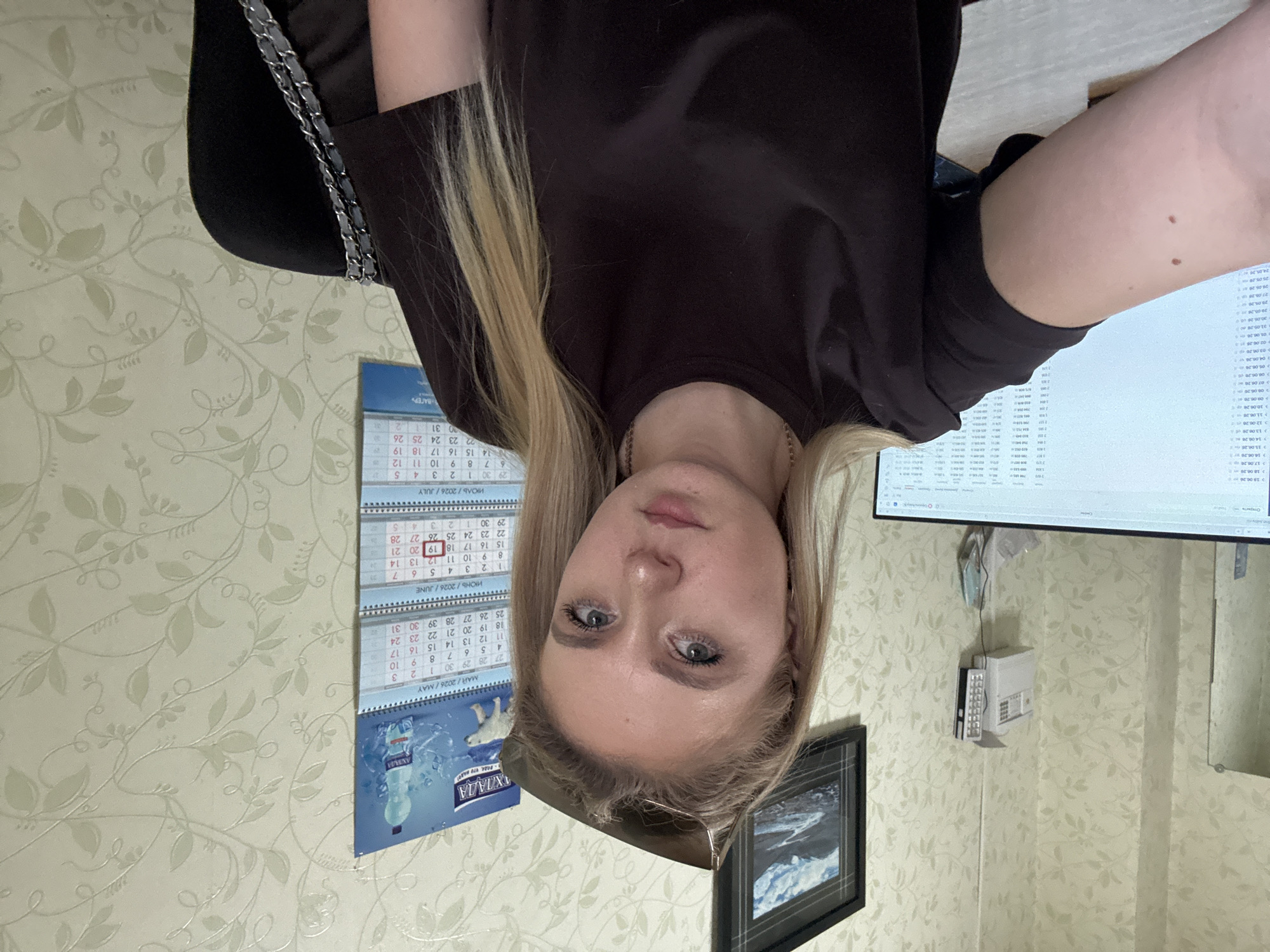 This is a photograph of a young woman with blonde hair, looking at the camera. She is wearing a dark brown shirt. She appears to be indoors, possibly at home or in an office, given the presence of calendars on the wall and a computer monitor. The calendars suggest the year 2026. There is also a framed picture of a snowy landscape on the wall. The lighting suggests it could be daytime. The woman's expression is neutral. The overall scene is casual and domestic. Visible text includes "АХЛАДА", "МАЙ/2026/MAY", "ИЮНЬ/2026/JUNE", "ИЮЛЬ / 2026/JULY", and "ВАГЕРЬ".