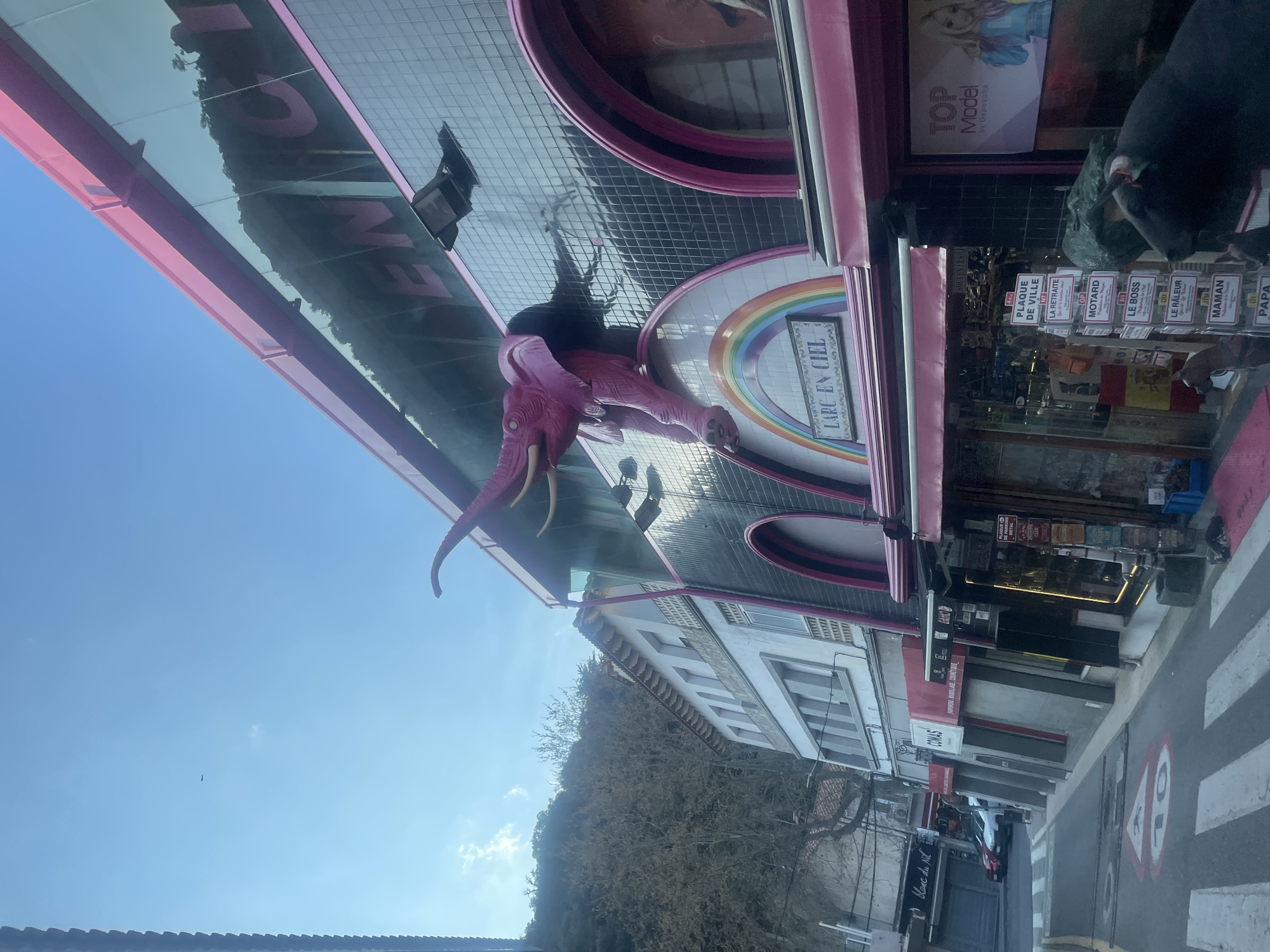 The image depicts a street scene in Le Perthus, France, under a clear blue sky with sparse clouds. Dominating the right side of the frame is a multi-story commercial building featuring a distinctive pink and black facade. A large, pink elephant sculpture with tusks protrudes from the second story, appearing to break through a black tiled wall segment. Below the elephant, a semi-circular arch decorated with a rainbow motif bears the sign "L'ARC EN CIEL."
The upper section of this building incorporates reflective glass panels, reflecting the sky and a distant, tree-covered hillside. The ground floor houses storefronts with pink awnings. One window displays "TOP MODEL by Despacha," while an adjacent souvenir shop displays various items and a stand outside with numerous plaques listing terms such as "PLAQUE DE VILLE," "LA RETRAITE," "MOTARD," "LE BOSS," "LE RÂLEUR," "MAMAN," and "PAPA." Partially visible in the immediate foreground, a dark-colored statue, possibly a bull, stands on the pink-paved sidewalk.
To the left of this prominent building, a white, multi-story building with a tiled roof and multiple windows is visible. Further down the street, other storefronts, including "Blanc du Nil" and "COMAS" with a red awning, line the road. The street itself is asphalt, featuring white pedestrian crossing stripes and a yellow line. A red triangular road marking with a "10" and a pedestrian symbol is painted on the pavement, indicating a speed zone. Several vehicles, including red and white cars, are parked further along the street. A steep, sparsely vegetated hillside rises in the background behind the buildings on the left. Utility poles and wires are present on the left side of the street.