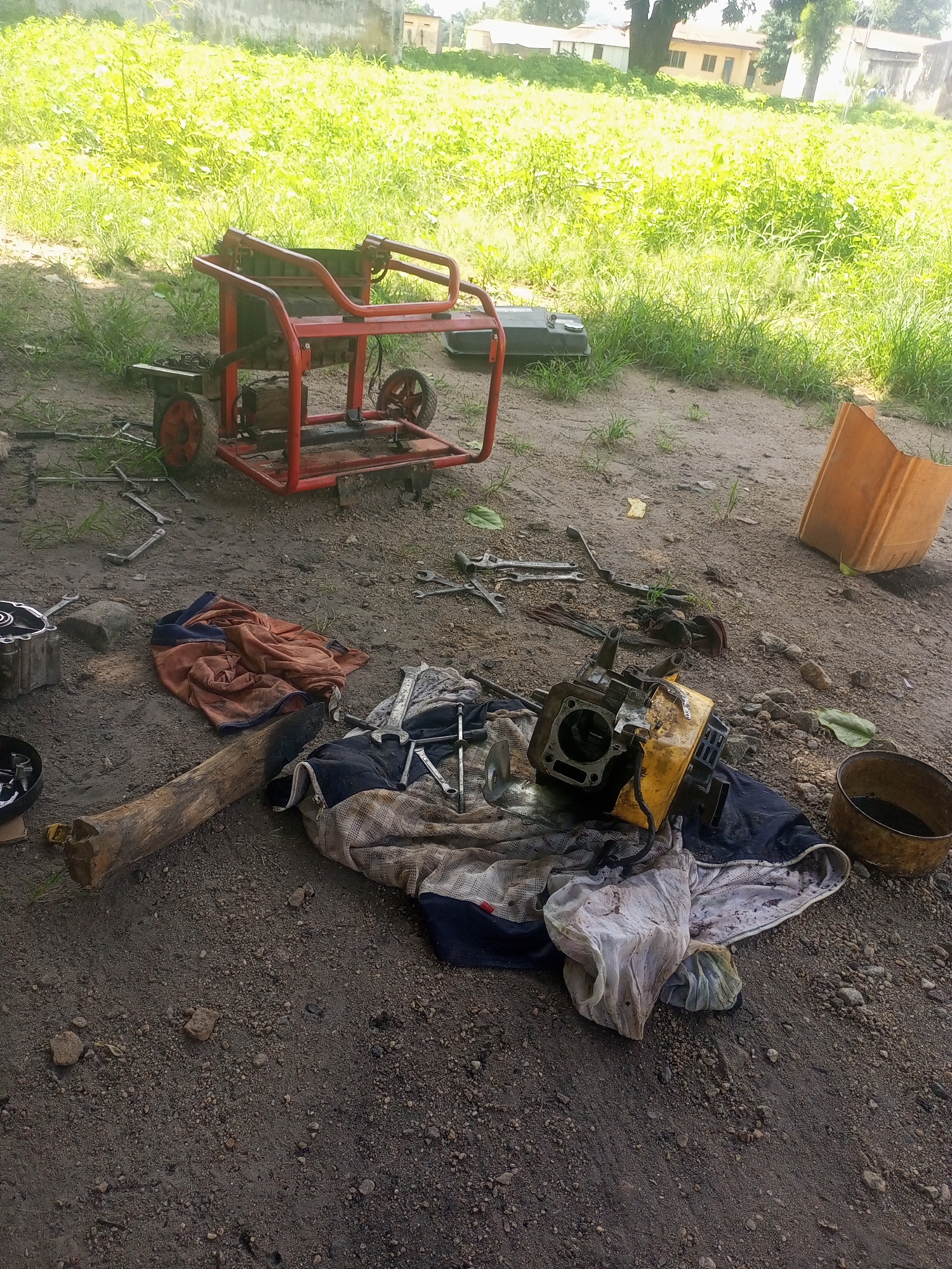 The image depicts an outdoor scene, likely a repair or workshop area, located in Serti, Nigeria. In the foreground, a red generator with two wheels is positioned on the dusty ground. Scattered around it are various tools, including wrenches and what appears to be engine parts. A pile of disassembled engine components, including a cylinder head and yellow casing, lies on the ground, covered by fabric. A wooden log and an orange plastic container are also visible. The background shows dense green vegetation and some buildings, suggesting a rural or semi-urban setting. The ground is a mixture of dirt and small stones. The lighting indicates it is daytime.