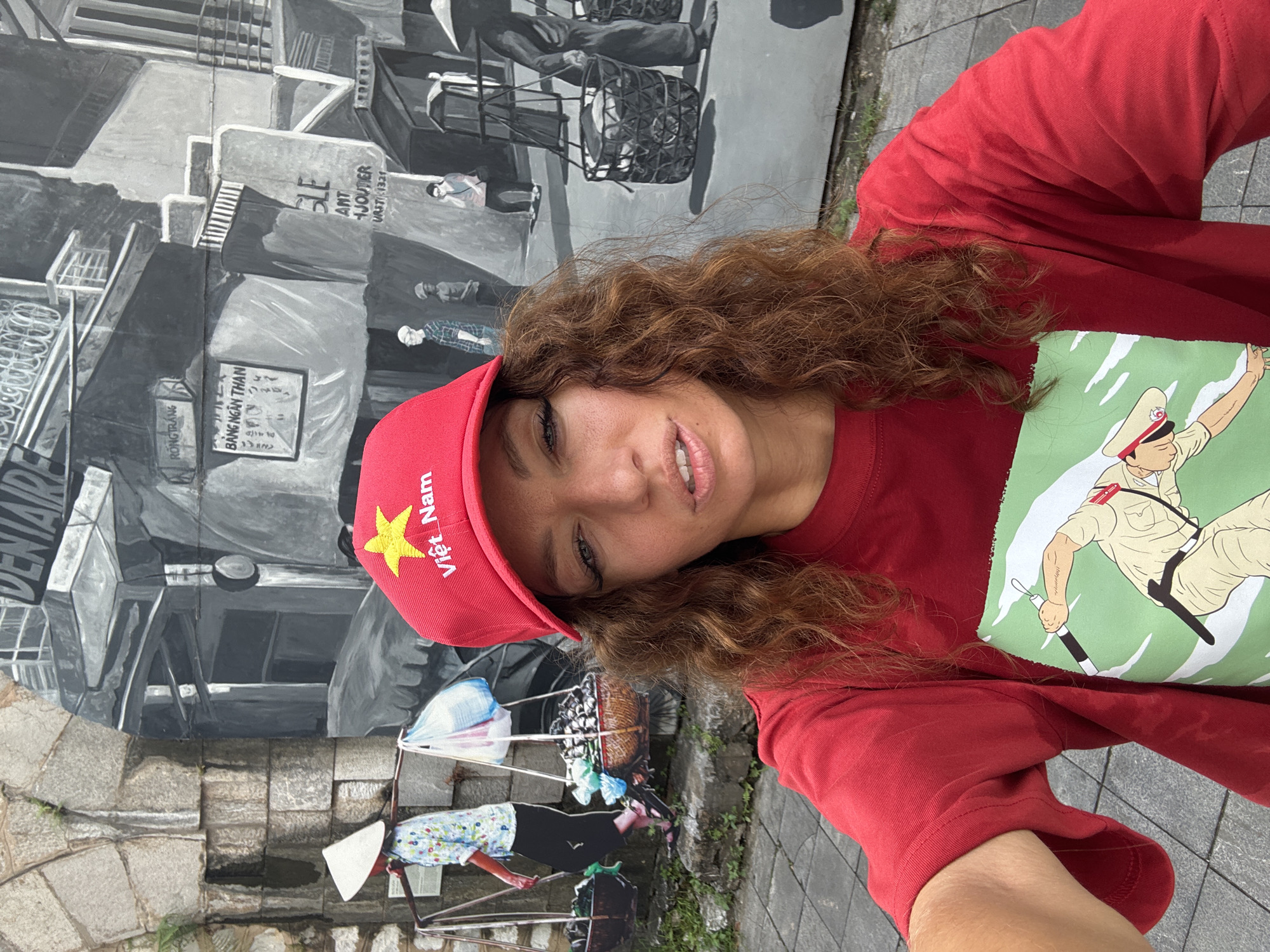 A woman with brown curly hair wears a red baseball cap with a gold star and the text "Việt Nam," and a red t-shirt featuring a cartoon illustration of a police officer. She is taking a selfie. The background is a mural painted on a wall. The mural is in black and white, depicting a street scene with people transporting goods in traditional Vietnamese style. In the lower part of the mural, a woman is carrying goods on a shoulder pole, while another scene above shows people riding rickshaws or tricycles. In the background of the mural, there are buildings and signs with Vietnamese text. The ground is paved with gray stone blocks.