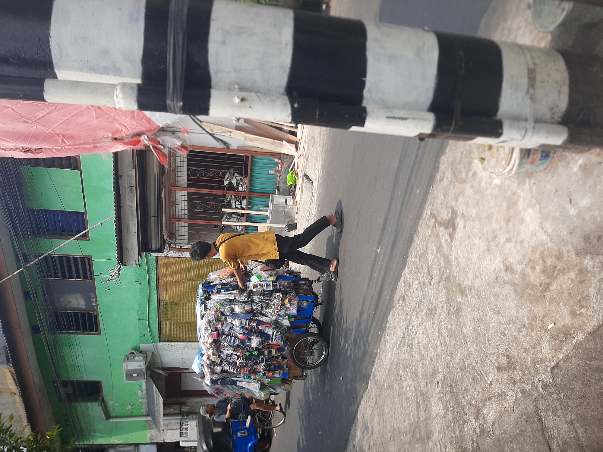 In the city of Central Jakarta, Indonesia, a man pushes a cart laden with various merchandise, including what appear to be small electronic accessories and keychains. The cart is a two-wheeled structure with a larger wheel at the back and smaller wheels at the front. The items on the cart are displayed in clear plastic packaging, creating a dense and colorful assortment.
The man, wearing a yellow t-shirt and dark pants, is walking on an asphalt road, pushing the cart with his right hand. He is wearing sandals. In the background, a blue motorized tricycle is visible, with a person sitting on it, looking towards the man with the cart.
The environment suggests a street scene. Buildings line the street, one of which is painted green and shows signs of weathering. There are windows with metal grills and an air conditioning unit mounted on the wall. Another building has a corrugated metal roof and a doorway with an orange gate. A tall, black and white striped pole stands prominently in the foreground, partially obscuring the view of the street. The ground is a mix of concrete and asphalt, with some debris scattered around. The overall impression is that of a busy urban environment during daytime.