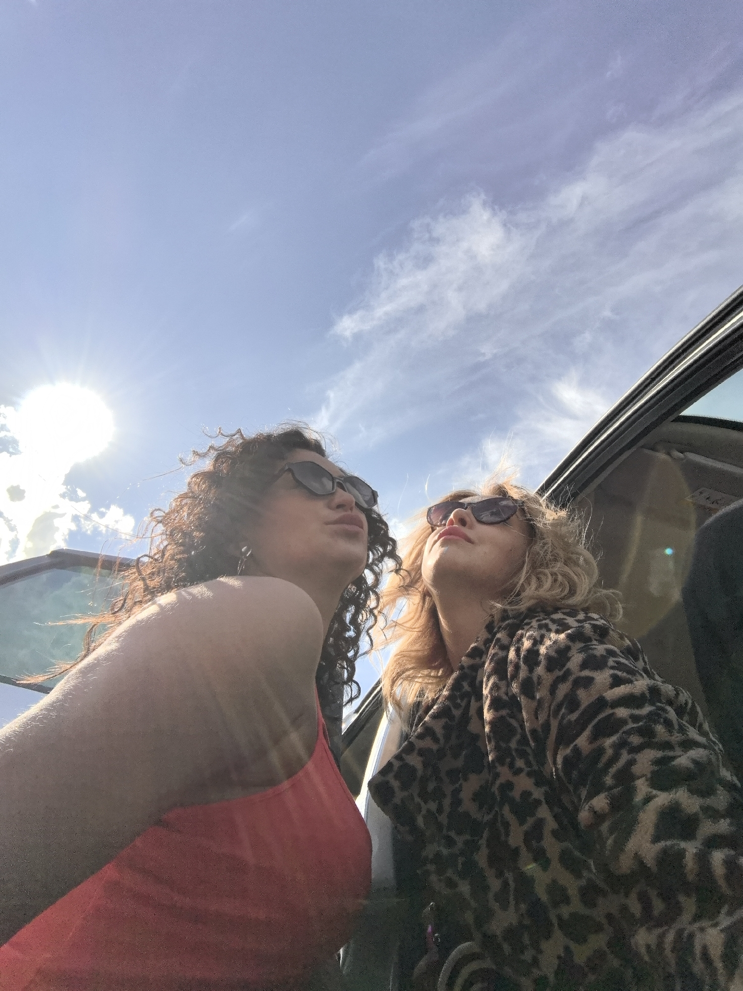 Two women are captured in a low-angle shot, looking upwards towards a bright, sunny sky from what appears to be a car, possibly a convertible or a vehicle with a sunroof, in Chaikovsky, Russia.
The woman on the left has dark, voluminous curly hair and wears dark sunglasses, partially obscuring her face. She is dressed in an orange or coral-colored sleeveless top, and her arm, which is closer to the camera, is visible. Her lips are slightly puckered, suggesting a playful or duck-face expression. Intense sunlight creates a bright lens flare in the upper left quadrant of the image, illuminating her from behind and above.
The woman on the right has blonde, curly hair and also wears dark sunglasses. She is clad in a distinctive brown and black leopard print jacket or coat. Her gaze is directed upwards, and her expression seems relaxed, enjoying the sun. She is positioned further into the vehicle than the first woman.
The sky overhead is a clear, light blue with scattered wispy white clouds. The sun is high, indicating it is likely midday or early afternoon, given the intensity of the light. No specific location cues other than the provided context of Chaikovsky, Russia, are visible. There is no visible text in the image. The overall mood suggests a carefree moment of enjoyment, possibly during a drive.