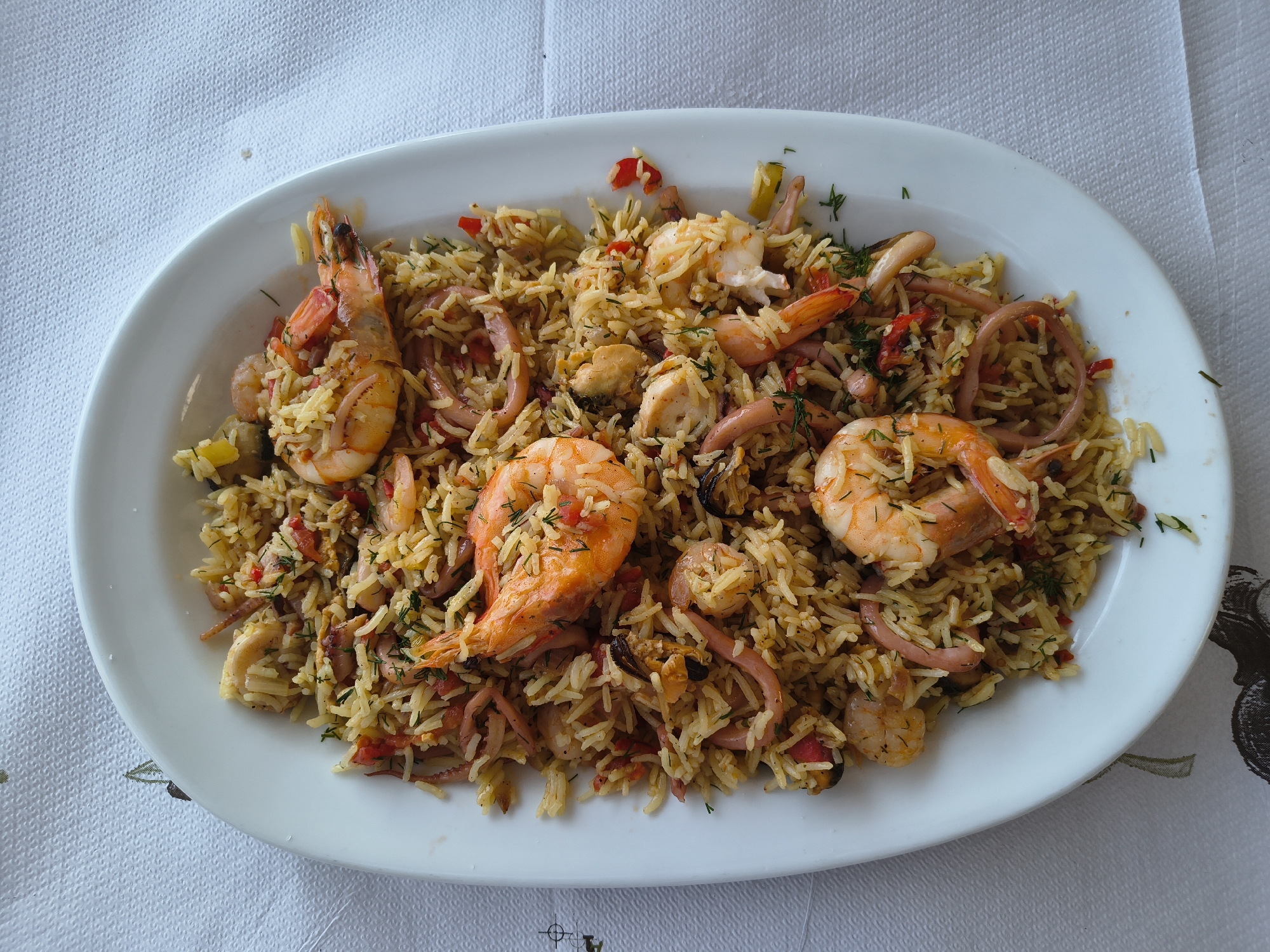 A high-angle, close-up shot shows a white oval platter filled with seafood rice served on a white tablecloth. The platter is positioned slightly off-center to the left. The rice is a golden-yellow color and appears to be mixed with small pieces of red pepper, green herbs, and possibly some other vegetables. Several large, pink shrimp are prominently displayed on top of the rice, with their tails pointing in various directions. Visible amongst the rice and shrimp are rings of squid and some mussels. The tablecloth has a subtle texture and a faint pattern, possibly floral, along the bottom edge. The scene is well-lit, and the focus is sharp on the food. The location is identified as Limenaria, Greece.