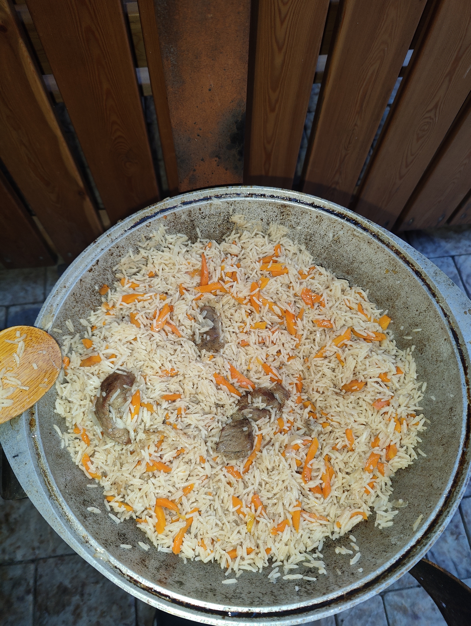 A top-down, medium shot captures a large, cast-iron kazan filled with pilaf, a traditional rice dish. The pilaf consists of long-grain rice, julienned carrots, and chunks of meat, likely lamb or beef. A wooden spoon with rice is visible on the left side of the kazan. The kazan itself is placed on what appears to be a tiled surface, possibly an outdoor patio. In the background, a wooden fence with vertical planks and a reddish-brown hue is present. The lighting suggests it is daytime, with natural light illuminating the scene. The context of Izberbash, Russia, implies this pilaf might be prepared for a gathering or a meal shared outdoors. The scene is static and focuses on the food preparation.