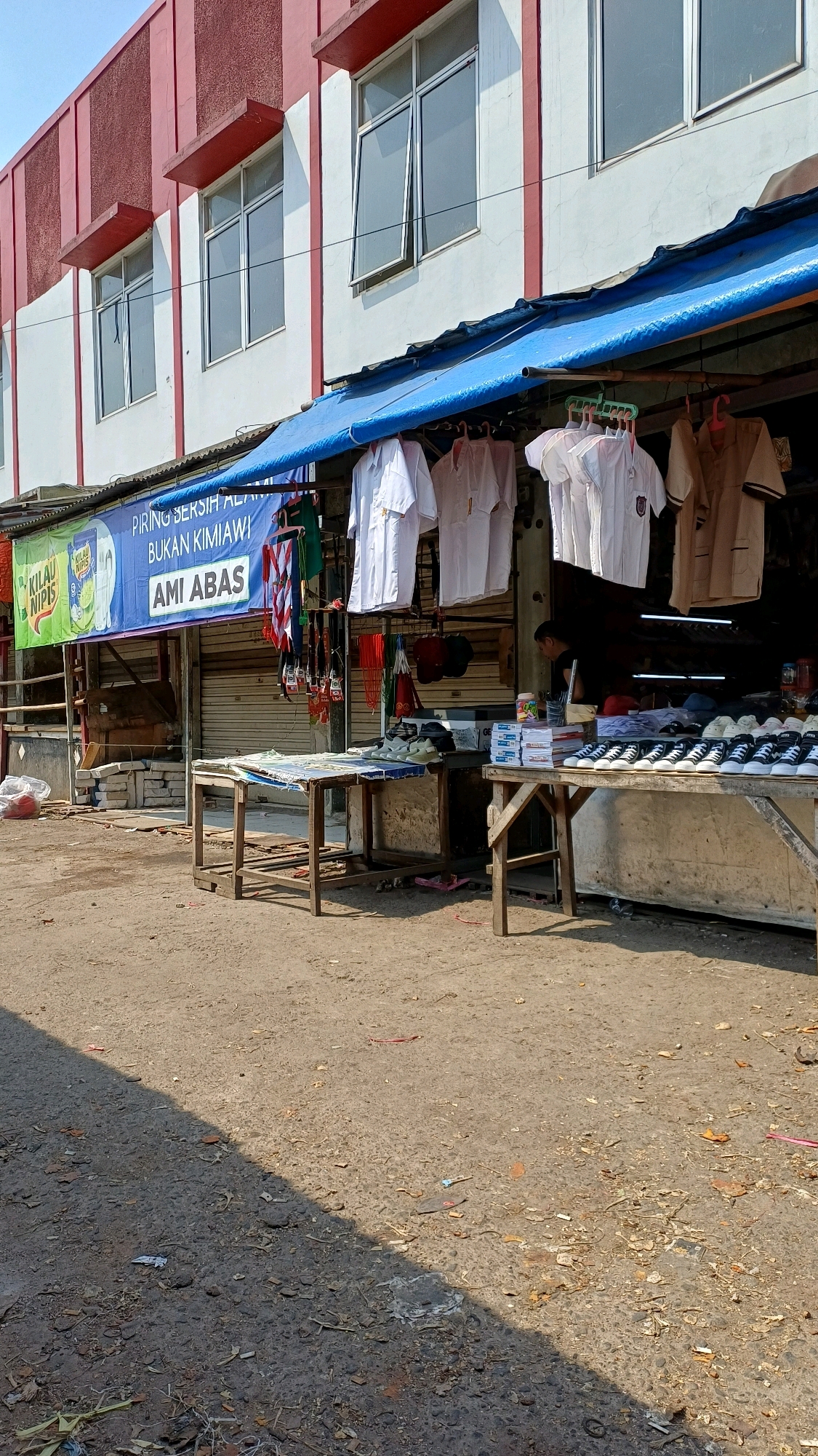 The image depicts a street-level view of a small storefront in Citeureup, Indonesia. The building facade is white with red trim and several windows. A blue awning extends from the building over a sidewalk, under which various goods are displayed for sale. White shirts, possibly school uniforms, and other garments are hung from hangers. In front of the storefront, two wooden tables are set up. One table displays stacks of books and what appear to be shoes. The other table has a collection of sneakers neatly arranged. The ground in front of the tables is a dirt and gravel surface, littered with debris. A man is partially visible in the background, inside the storefront. The overall scene suggests a small business operating outdoors.