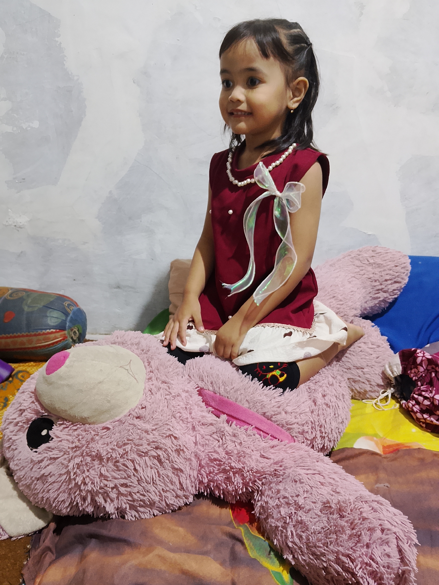 A young girl, appearing to be of Southeast Asian descent, sits atop a large, fluffy pink stuffed animal, likely a rabbit, in what appears to be a bedroom. The girl is wearing a maroon sleeveless dress with a white lace trim at the hem and a pearl necklace. A sheer ribbon is tied in a bow on the front of her dress. She has dark hair styled in braids and is looking to her right, with a slight smile. The stuffed animal is positioned on a bed, covered with a patterned duvet. In the background, a wall with a textured, mottled grey and white surface is visible. To the left of the stuffed animal, a cylindrical pillow with a colorful, abstract pattern rests on the bed. To the right, a blue pillow and a piece of fabric with a floral pattern are also present. The lighting in the room appears to be artificial, casting soft shadows. The setting is likely residential, with the presence of bedding and toys suggesting a domestic environment. This scene is located in Tanggungan, Indonesia.