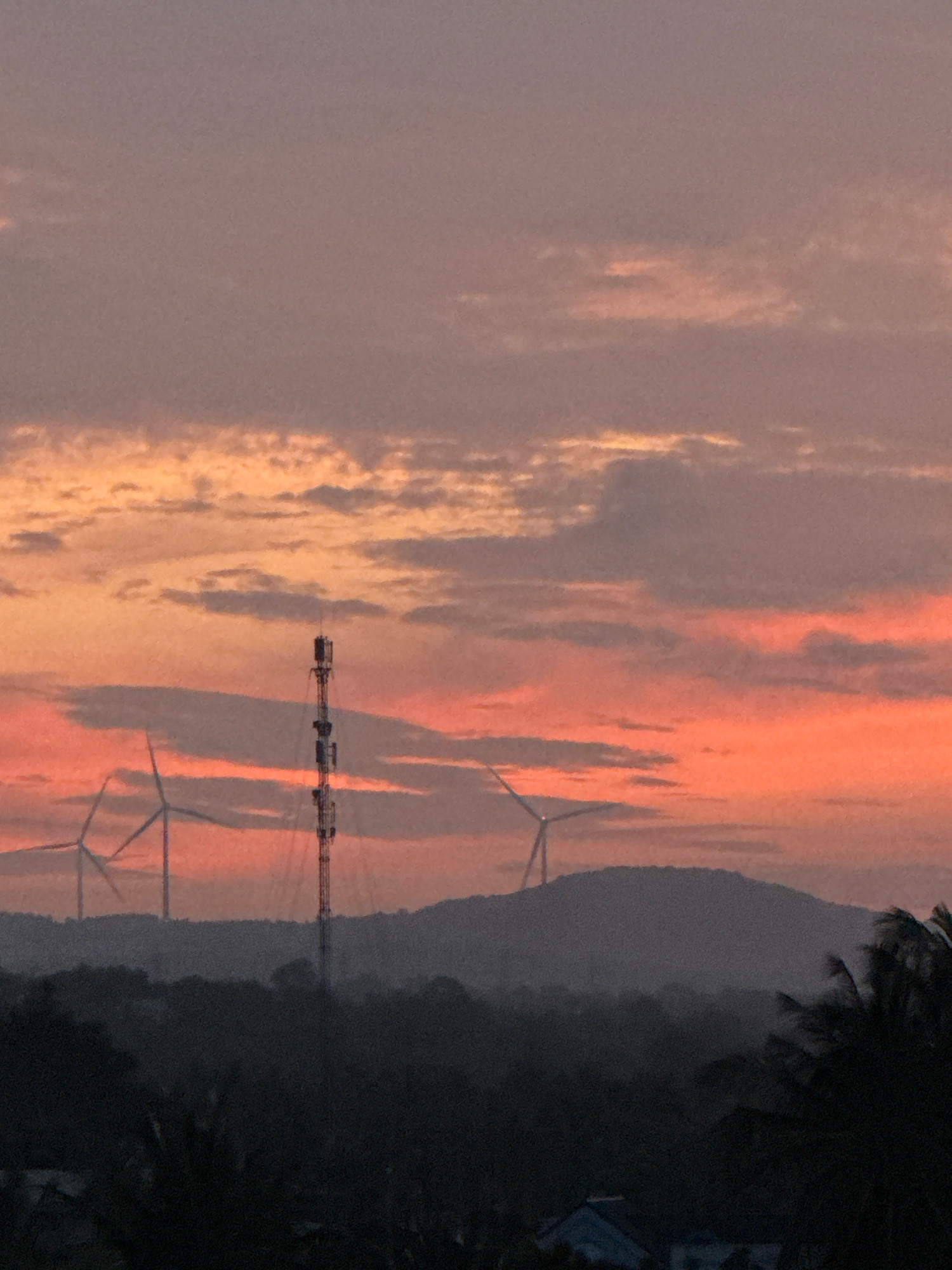 This is a wide-angle, high-angle shot of a sunset in Mui Ne, Vietnam. The sky is a gradient of warm colors, ranging from deep orange to pink and purple, with scattered clouds. In the middle ground, a tall, slender tower, likely a telecommunications tower, stands prominently. To the left and right of the tower, two wind turbines with their blades outstretched are visible, silhouetted against the sky. They are situated on rolling hills that are partially obscured by atmospheric haze. The foreground consists of dark, silhouetted foliage, possibly palm trees, and the faint outline of buildings. The overall scene conveys a sense of natural beauty and technological infrastructure coexisting in a coastal landscape.