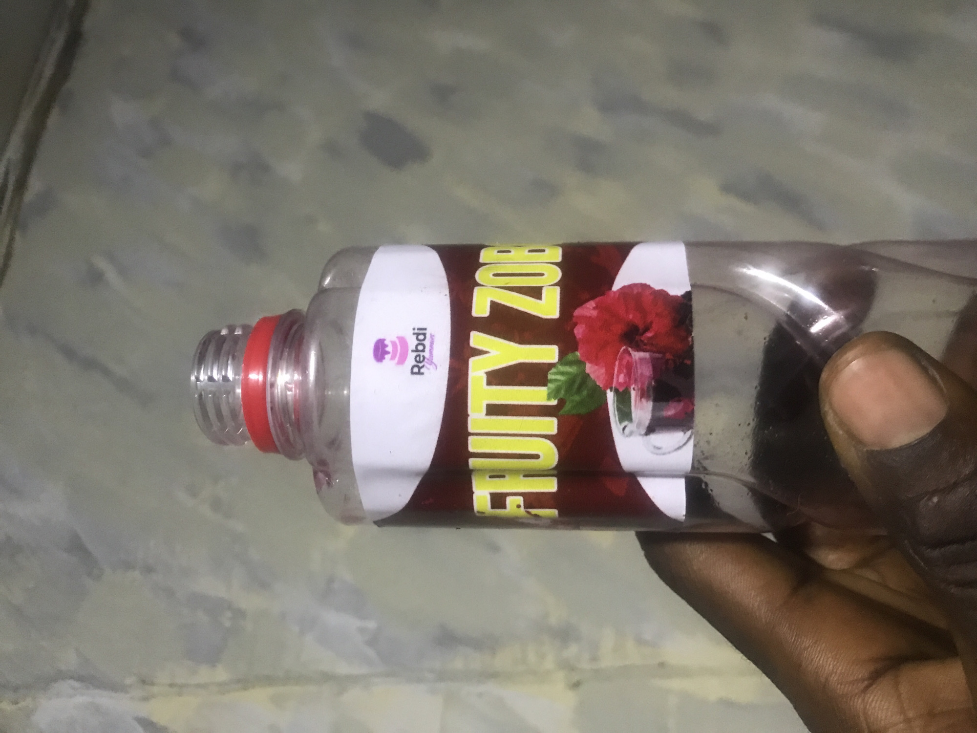 A close-up, horizontal shot shows a dark-skinned hand holding a clear plastic bottle against a muted, patterned background. The bottle has a red screw cap and a prominent label. The label features a large, dark red banner with "FRUITY ZOB" written in bright yellow, bold capital letters. To the right of this text, an illustration depicts a vibrant red hibiscus flower alongside a glass filled with a dark red liquid, suggesting the drink is hibiscus-based. To the left of "FRUITY ZOB," on a white section of the label, there is a small logo that includes the word "Rebdi" with smaller text below it, possibly "yummy," and an icon resembling a cupcake or baked good. The bottle contains a dark liquid, likely a beverage. The background appears to be a light gray or greenish wall or surface with darker, irregular smudges or patterns, providing a simple and somewhat indistinct setting. The lighting is soft and ambient, without direct bright light. The image focuses on the product and the hand holding it, presenting the bottle's design and contents. No specific time of day or outdoor weather conditions are discernible, and no strong emotions are conveyed. The context provided indicates the location is Rafin Par, Nigeria.