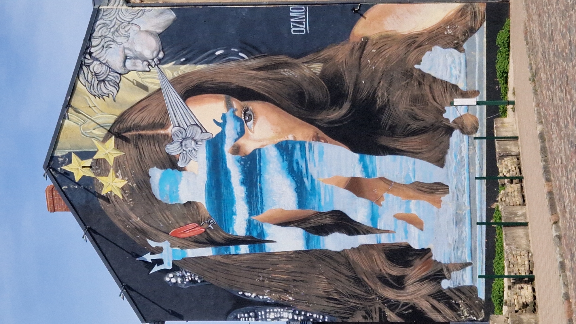 The image shows a mural on the side of a building in Liepaja, Latvia. The mural depicts a large, fragmented silhouette of a woman, revealing a seascape of waves and sky within the silhouette. The woman has long, brown hair and is adorned with a crown of three stars and a metallic flower on her forehead. To the right of the woman's face, a profile of a classical bust is depicted blowing air, which appears to be interacting with the woman's hair. The building itself has a sloped roof and a brick chimney. In the foreground, there is a cobblestone street, a low wall with some greenery, and a few metal posts. The sky is a clear blue. The mural is signed "OZMO".