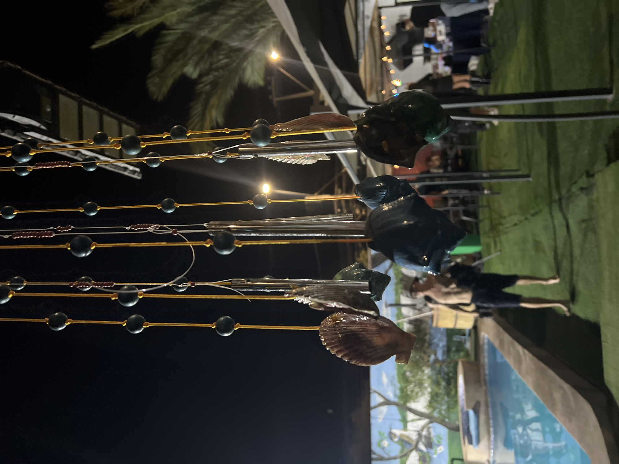 The image shows outdoor lighting with suspended decorations. The decorations consist of strings of beads interspersed with seashells, and what appear to be dark green and black decorative pieces. A section of artificial green turf is visible in the background, along with people, tables, and what appear to be other decorations. A mural is partially visible. A portion of a pool is located in the lower right corner. Based on the objects in the image, it could be located in the city of Arica, Chile.