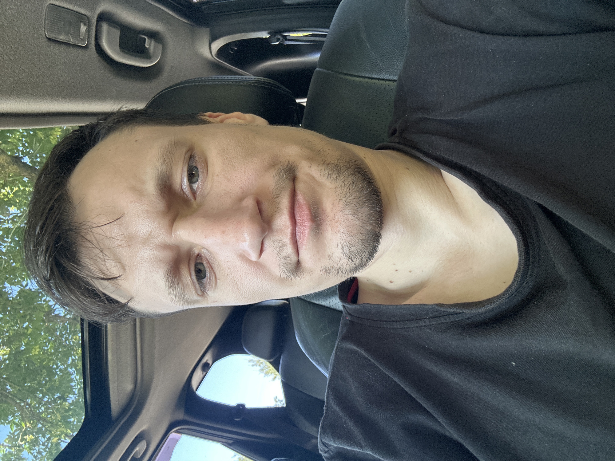 The image is a close-up, selfie-style photograph taken from inside a vehicle. The subject is a man with fair skin, dark hair, and a light beard, looking directly at the camera. He is wearing a black t-shirt. The interior of the car is visible, including a black leather headrest and seat behind him, a sunroof with trees and sky visible through it, and a portion of the dashboard. The scene appears to be outdoors, possibly in a rural or suburban setting, given the presence of trees and the natural lighting. There is no indication of the specific location within the city of Makiv, Ukraine.