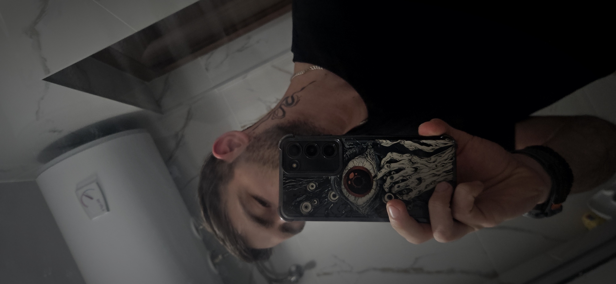 This image is an inverted mirror selfie, likely taken in a bathroom in Plovdiv, Bulgaria. A male figure is reflected, visible from the chest up, with his head tilted. He has short hair, a beard, and an earring on his visible ear. A stylized tattoo adorns his upper left arm/shoulder. He wears a dark (likely black) t-shirt and a dark bracelet or watch on his left wrist.
He holds a smartphone with his right hand, positioning it to capture the reflection. The phone features a multi-lens camera system on its back. Its case displays a distinctive, dark art design: a large, realistic eye with a prominent red iris is central, surrounded by multiple smaller eyes and several skeletal, reaching hands.
The background shows light-colored, marble-patterned tiles on the walls. To the left, a white cylindrical water heater (boiler) is reflected, featuring a control panel with a red indicator dial; however, specific text on the dial or panel is illegible. A chrome bathroom fixture is partially visible in the lower-central part of the reflection. The indoor setting suggests artificial lighting, making the time of day indeterminate. The person's expression is unreadable due to the angle and reflection.