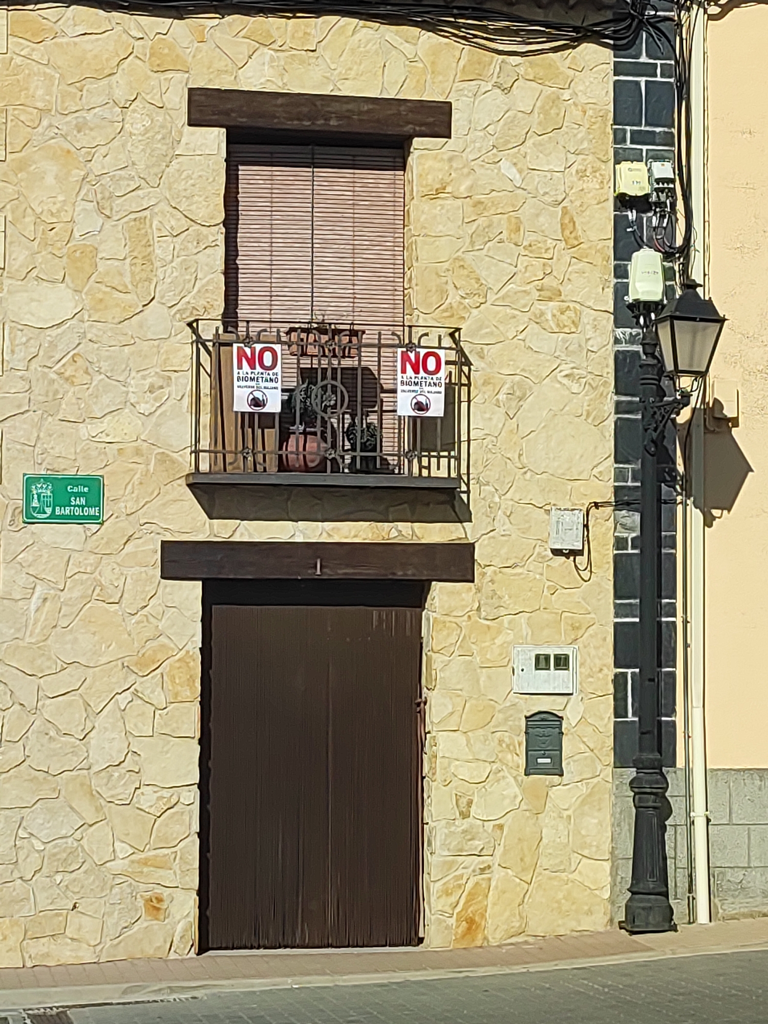 The image shows the facade of a building in Valverde Del Majano, Spain. The building has a stone exterior and a wooden door. There is a balcony with two signs that say "NO A LA PLANTA DE BIOMETANO" which translates to "NO TO THE BIOMETHANE PLANT". The signs also have a picture of a biogas digester crossed out. A street lamp stands on the right side of the building. A street sign on the left reads "Calle San Bartolomé". There are electrical boxes and a mailbox on the wall of the building. The scene appears to be an outdoor setting on a sunny day.