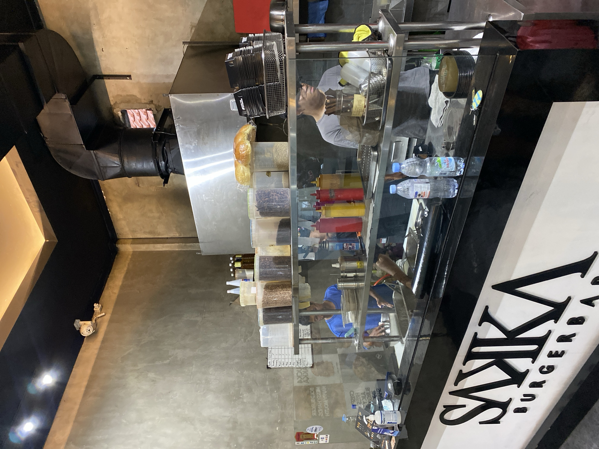 The image shows the interior of a burger bar in North Kuta, Indonesia. The burger bar has a modern industrial design with concrete walls and stainless steel kitchen equipment. In the foreground, a black counter with the "SAIKA BURGERBAR" logo is visible. Behind the counter, two employees are working in the kitchen. One employee, wearing a blue shirt, is seen preparing food. The other employee, wearing a grey t-shirt and glasses, is standing near the cooking area. Shelves above the counter are stocked with various ingredients in transparent containers, including what appear to be spices, grains, and buns. Condiment bottles are also visible on the shelves. The overall atmosphere suggests a busy food establishment.