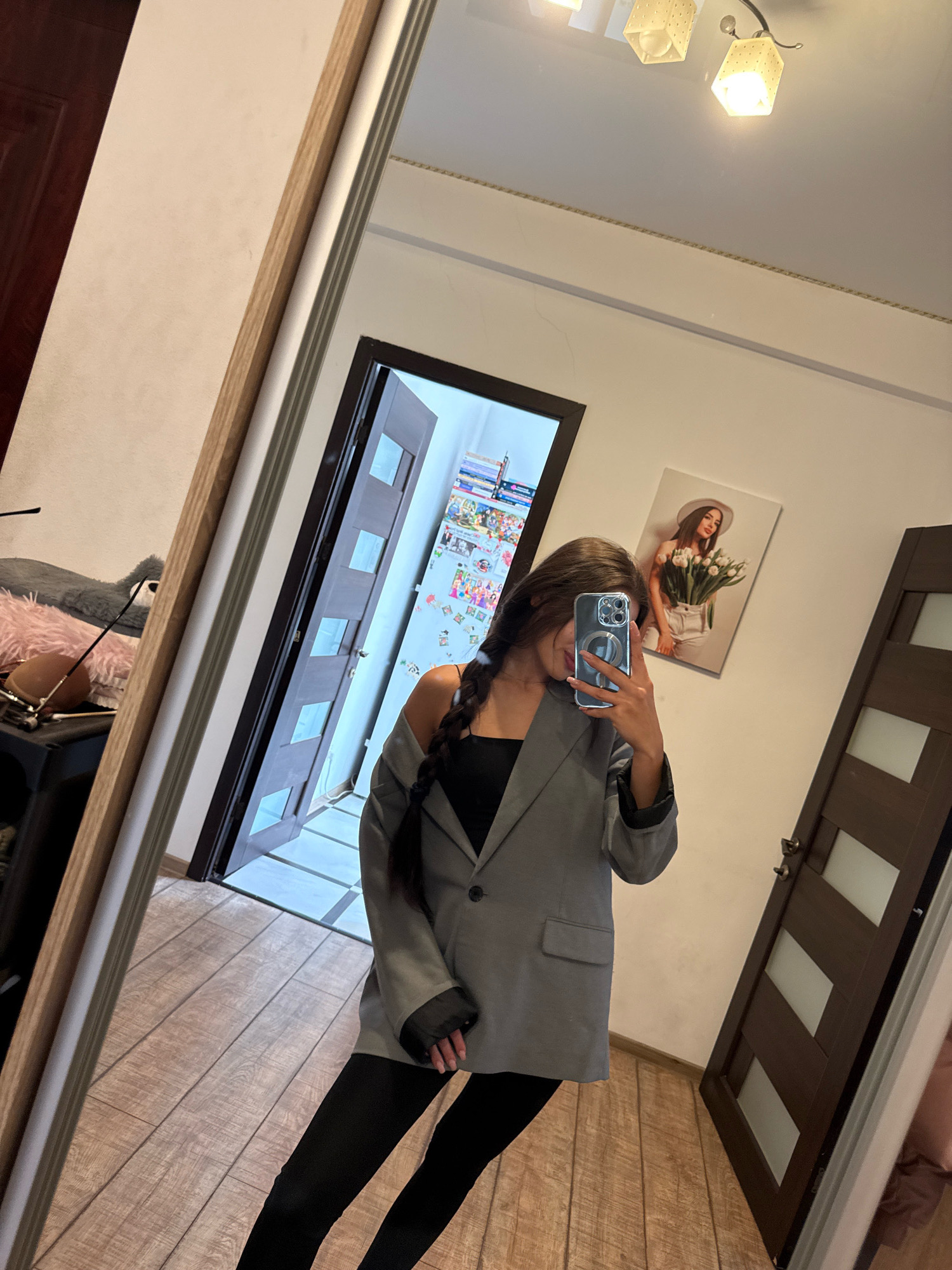 The image depicts a young woman taking a mirror selfie in what appears to be an indoor setting, possibly a hallway or living room. She is wearing a grey blazer over a black top and black leggings. Her hair is long and dark, styled in a braid. She is holding a smartphone to take the picture.
In the background, a large mirror reflects the scene. To the left of the mirror, part of a wooden doorframe and a doorway leading to another room are visible. The room beyond the doorway appears to be decorated with various items, including what looks like a calendar or posters on the wall.
To the right of the woman, another door with glass inserts is visible. On the wall between the doors, a framed picture of a woman holding a bouquet of flowers is displayed. The flooring is a light-colored wood laminate. The lighting appears to be artificial, coming from ceiling fixtures. The overall impression is that of a residential interior.
The user has indicated that the location is Chișinău, Moldova. Without additional information or context within the image itself, it is not possible to definitively confirm or deny this location. The scene is consistent with a typical residential interior found in many urban environments.