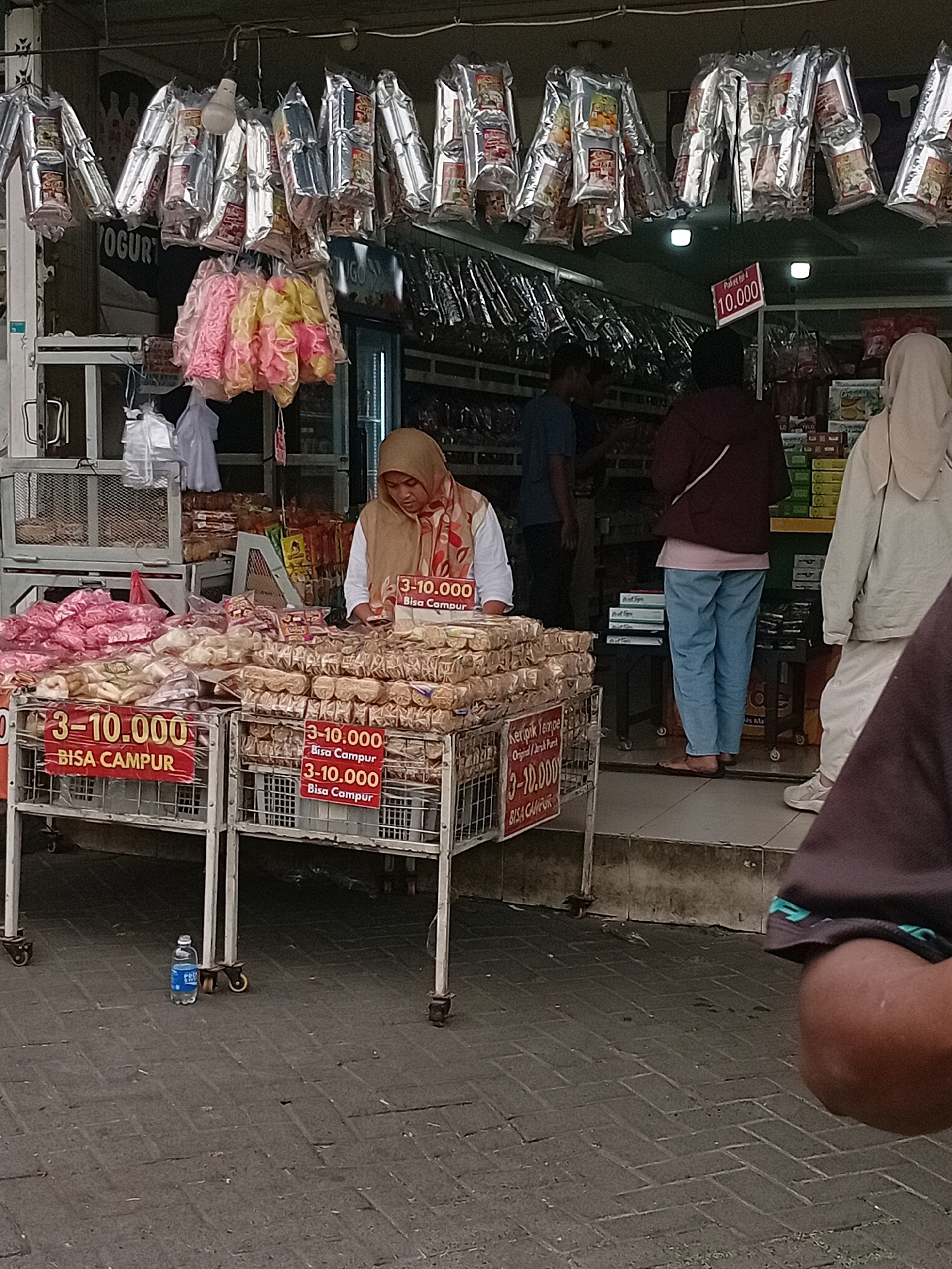 The image depicts an outdoor market stall in Lawang, Indonesia. A woman wearing a hijab and a patterned scarf is standing behind a display of packaged snacks. She appears to be tending to the stall, which is laden with various food items. Two tiered metal carts filled with individually wrapped snacks are positioned in front of the stall. Red signs with white lettering indicating "3-10.000" and "BISA CAMPUR" (meaning "can mix") are attached to the front of the carts. In the background, other market stalls and shoppers are visible. The ground is paved with interlocking bricks. A blue plastic water bottle is on the ground in front of one of the carts. The overall scene suggests a busy commercial environment where food items are being sold.