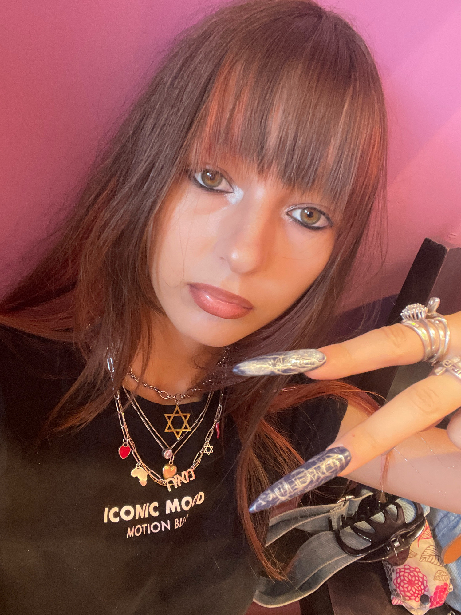 A young woman with long, dark brown hair and bangs with subtle red highlights is shown in a close-up, selfie-style shot. She is wearing a black t-shirt with white text that reads "ICONIC MOOD MOTION BL". She is adorned with multiple layered necklaces, including a prominent gold Star of David, a red heart charm, a pink heart charm, an outline of the continent of Africa, and a charm with Hebrew lettering. Her hands are also decorated with elaborate, long, pointed nails painted with a metallic blue and white marbled design. She wears several silver rings on her fingers, including one with a cluster of small stones. Her makeup features dramatic dark eyeliner and light-colored eyeshadow. The background is a plain, solid pink wall. The image does not contain any recognizable landmarks or contextual clues that would allow for the determination of a specific location within the city of Raanana, Israel.