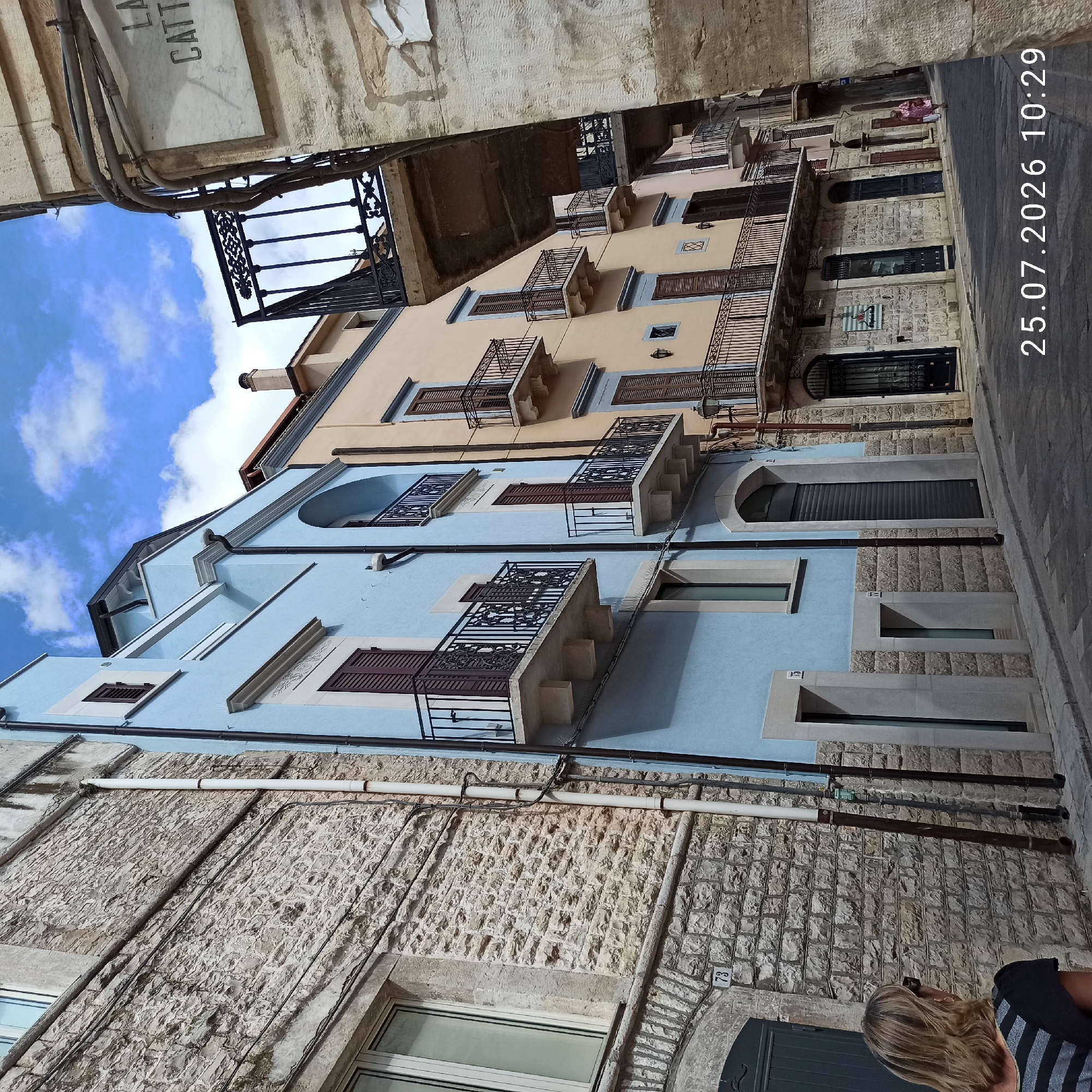 The image shows a street scene in the historic center of Ruvo di Puglia, Italy, with a date and time overlay reading "25.07.2026 10:29". The scene features traditional stone buildings and brightly colored facades lining a narrow, paved street.
On the left, a building constructed with rough-hewn, pale stone dominates the foreground. It has a large window with a light-colored frame and a textured, almost rough surface on its facade. Beneath this window, a gray door is visible, marked with the number "73".
Adjacent to the stone building is a brightly painted light blue structure. This building has multiple windows with dark shutters and wrought-iron balconies with ornate railings, supported by stone corbels. The architectural style suggests a historical Italian setting.
Further down the street, a pale peach-colored building is visible, also featuring dark shutters and wrought-iron balconies. The buildings are tightly packed, creating a sense of an urban canyon.
In the background, a woman with blonde hair wearing sunglasses and a dark striped shirt is partially visible, looking towards the buildings. Further down the street, a person in a pink outfit is walking away from the camera.
The sky above is blue with scattered white clouds. The street is paved with cobblestones or similar material, and there are drains and utility lines visible along the buildings. A marble sign with black lettering, partially obscured by wires, is mounted on a stone structure on the right side of the image. The visible text on the sign appears to read "LA CATTI".
The overall impression is of a sunlit day in a picturesque Italian town, with a blend of ancient stone architecture and more recent, colorful renovations.