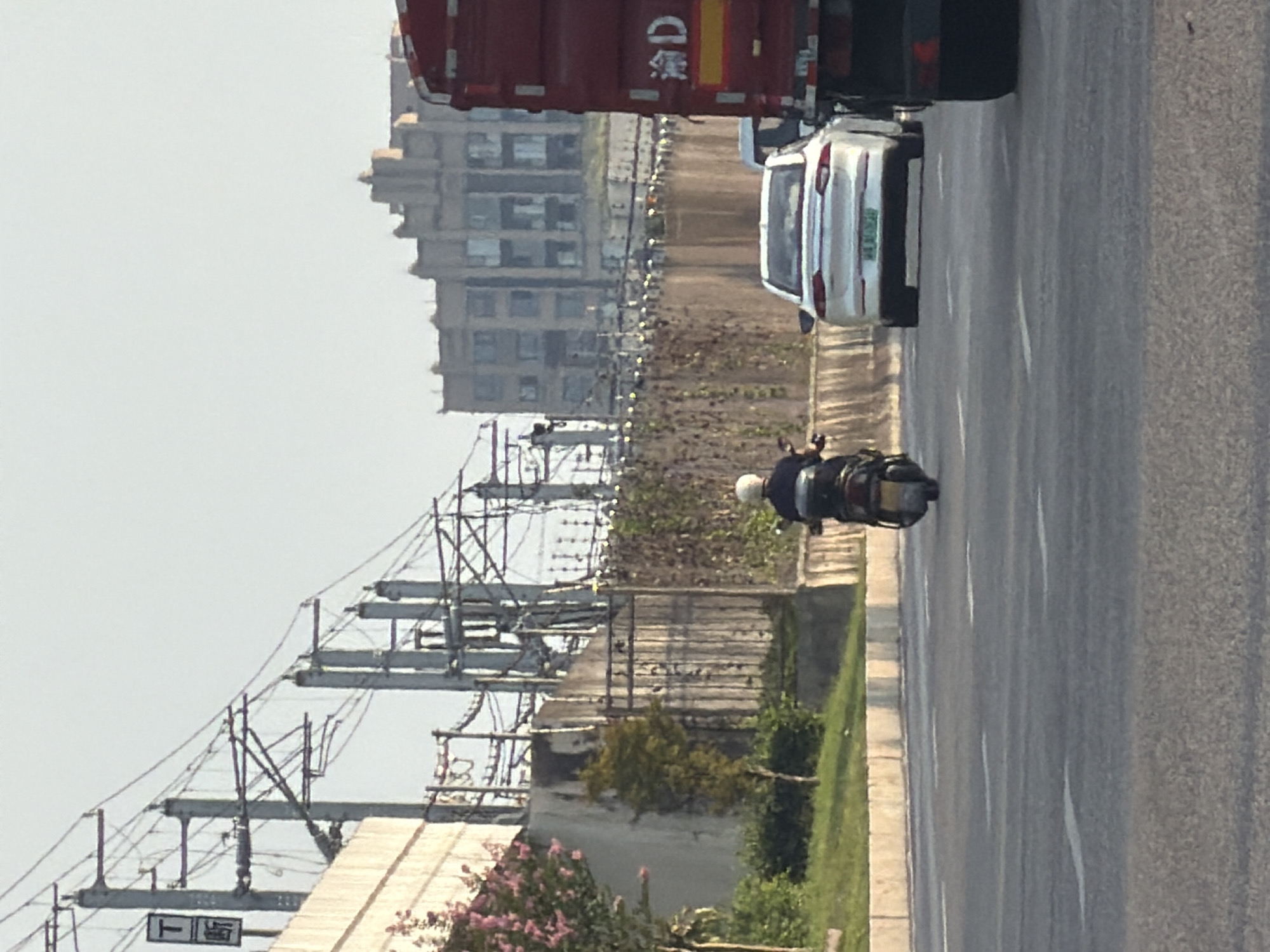 In Jiangjin, China, a low-angle, eye-level medium shot captures a street scene. The foreground features a paved road with white lane markings. In the middle ground, a person on a black scooter with a yellow license plate is traveling away from the camera, wearing a white helmet. To the right of the scooter, a white sedan with a green license plate is also moving away. Further to the right, the back of a large red truck is visible, with a yellow and red stripe on its rear bumper and the characters "冷 D" on its side. Beyond the road and vehicles, a concrete wall runs horizontally, topped with a fence. Behind the wall, a building with multiple floors and a complex architectural design stands against a pale sky, with visible overhead power lines and utility poles. Green grass and some trees are present on the left side of the scene, with a flowering tree in bloom. A concrete barrier runs along the edge of the road in the foreground.