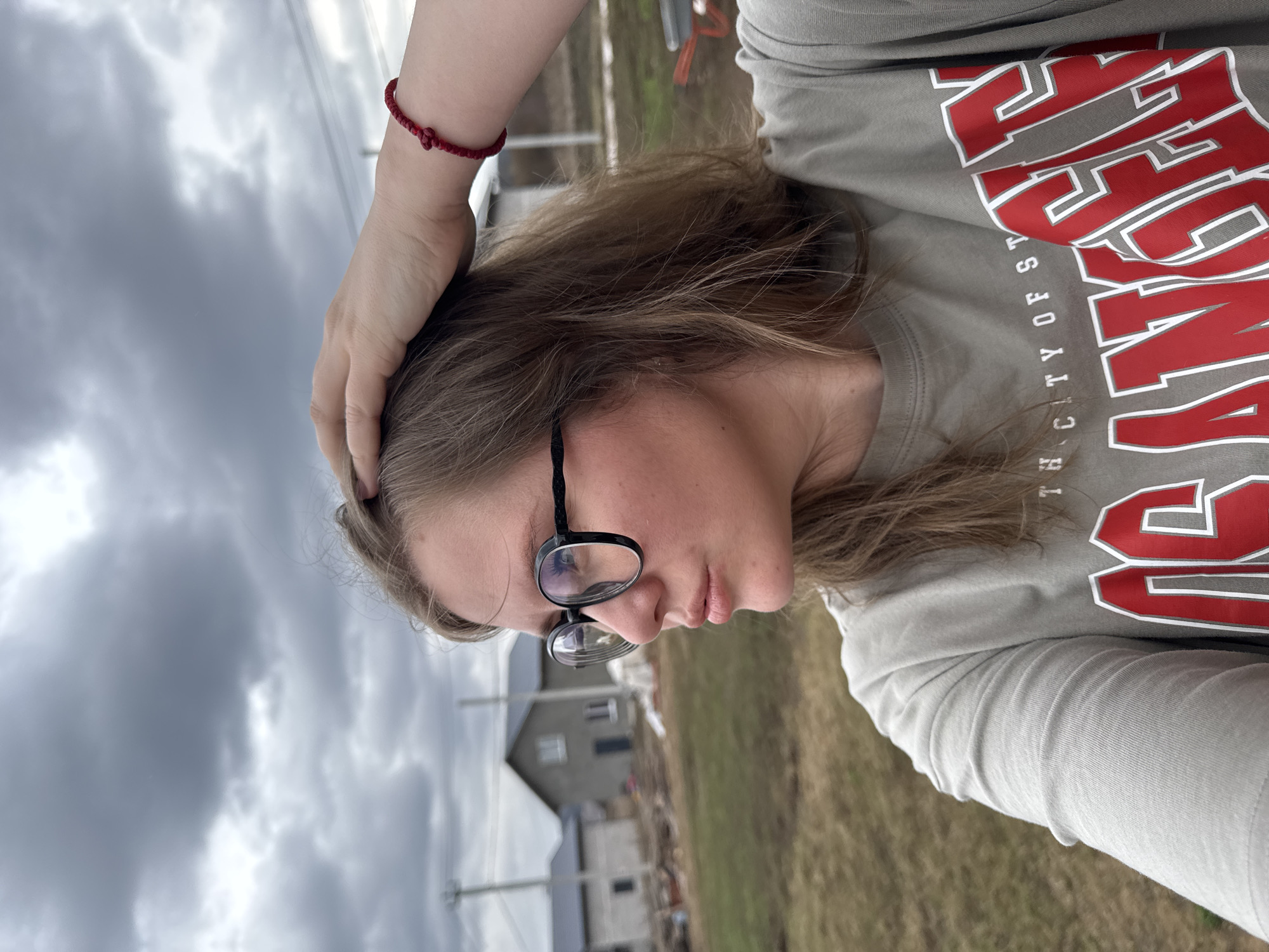A young person with light brown hair and glasses is shown in a selfie-style image. They wear a gray t-shirt with red lettering that reads "CITY OF ST..." (partially obscured). The person's left hand rests on their head, and a red bracelet is visible on their wrist. In the background is a gray house, partially obscured by vegetation, and a cloudy sky. The location is the city of Gornyi in the Dvurechenskoe S/P area of Russia. The ground is covered in sparse, dry grass.