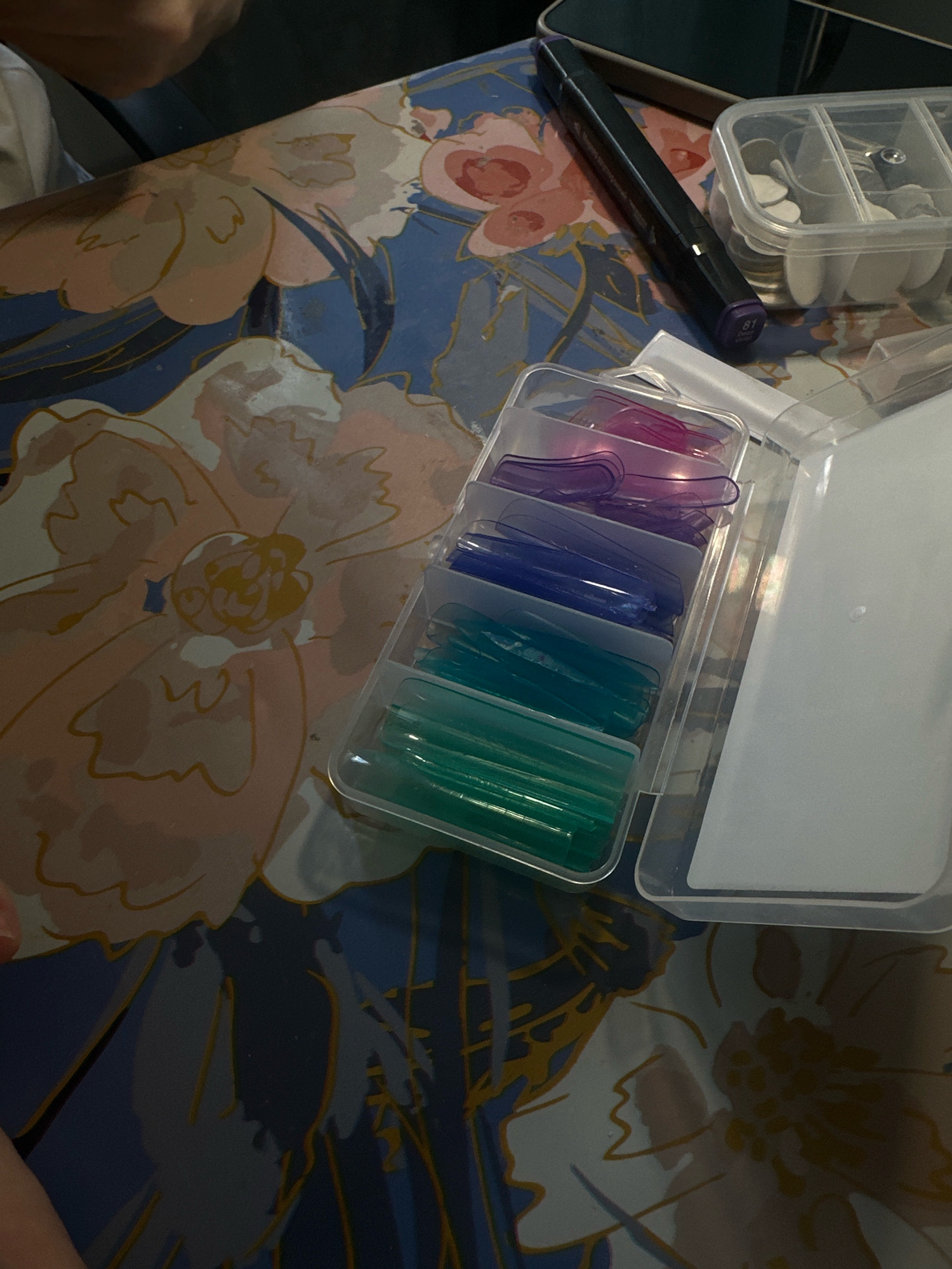 In an indoor environment, a table is covered with a blue tablecloth featuring a floral pattern of pink and yellow flowers. On the table, a black marker with a purple cap and the number "81" visible is placed next to a clear plastic container holding small white objects, possibly nail tips. To the left, a clear plastic case contains multiple rows of colored nail tips, ranging from pink and purple to blue and green. The overall scene appears to be a workspace or a collection of nail art supplies. The image is taken from a slightly elevated angle, showing a portion of a person's arm and hand in the upper left corner, and a person's thumb and index finger in the lower left corner.